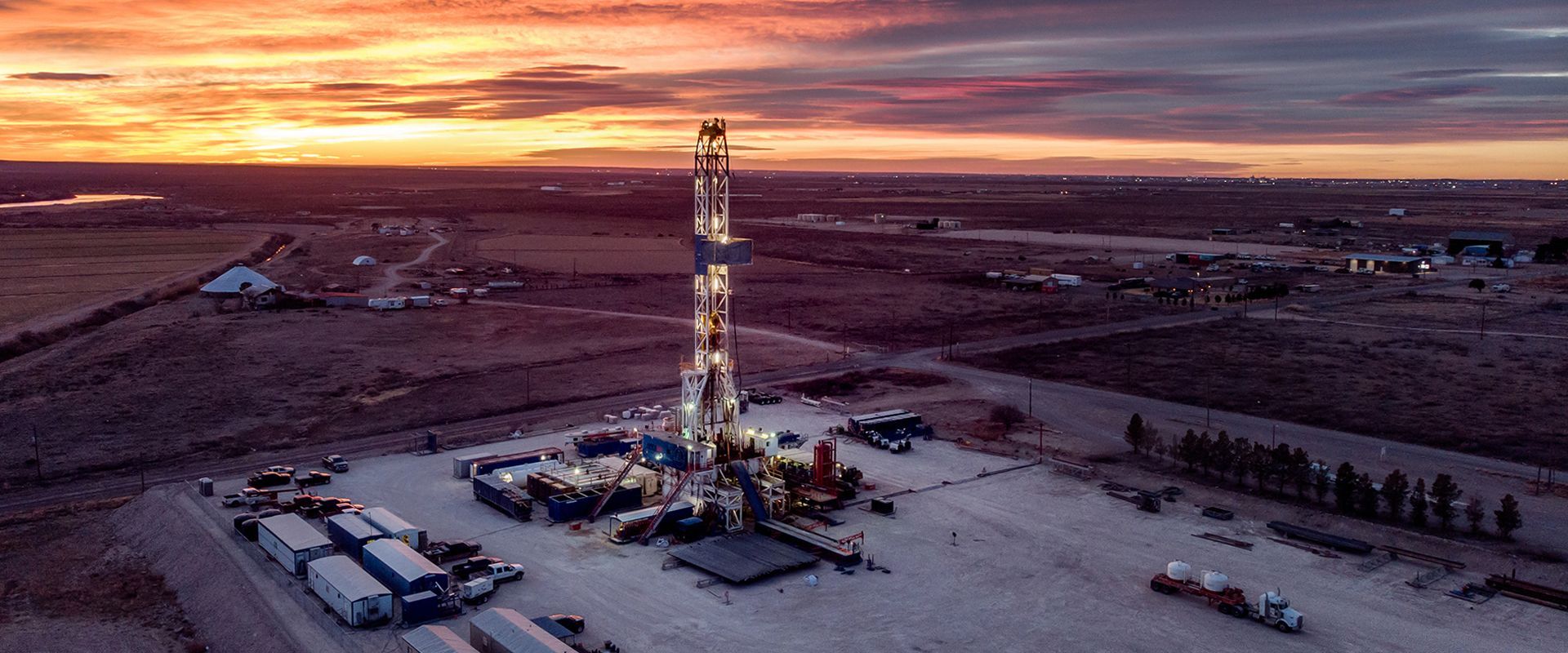 Oil drilling rig at sunset in a vast desert, with trucks and equipment around the site.