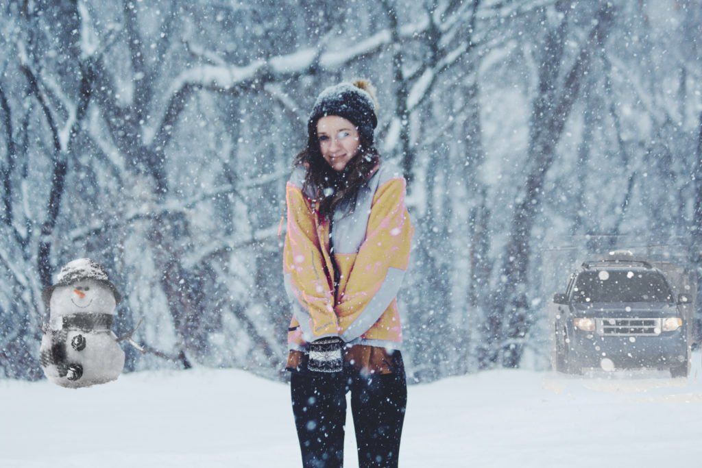 A woman is standing in the snow in front of a snowman and a car.