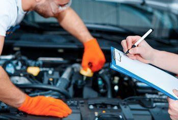 A man is working on a car engine while a woman writes on a clipboard.