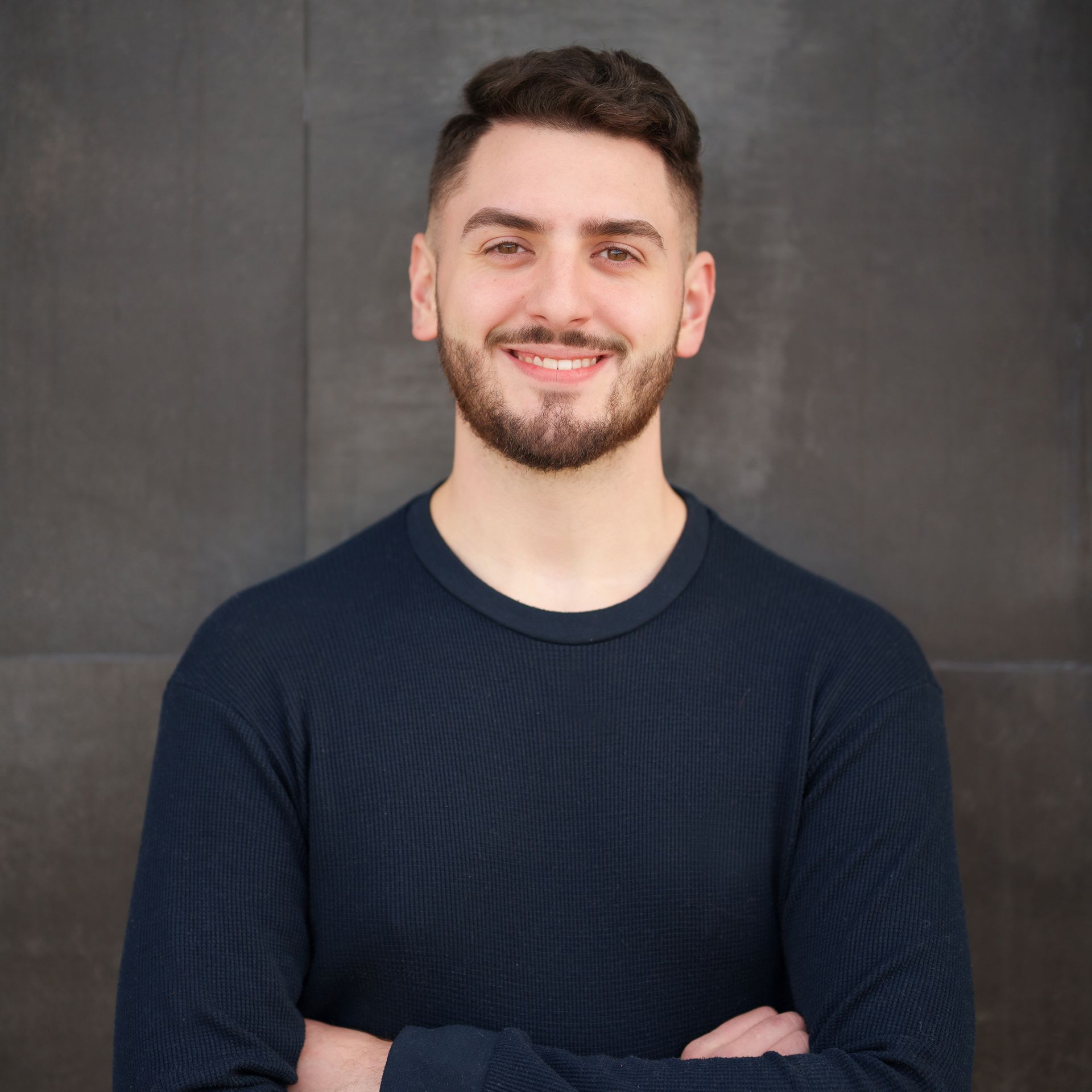 A man with a beard wearing a blue sweater is smiling with his arms crossed