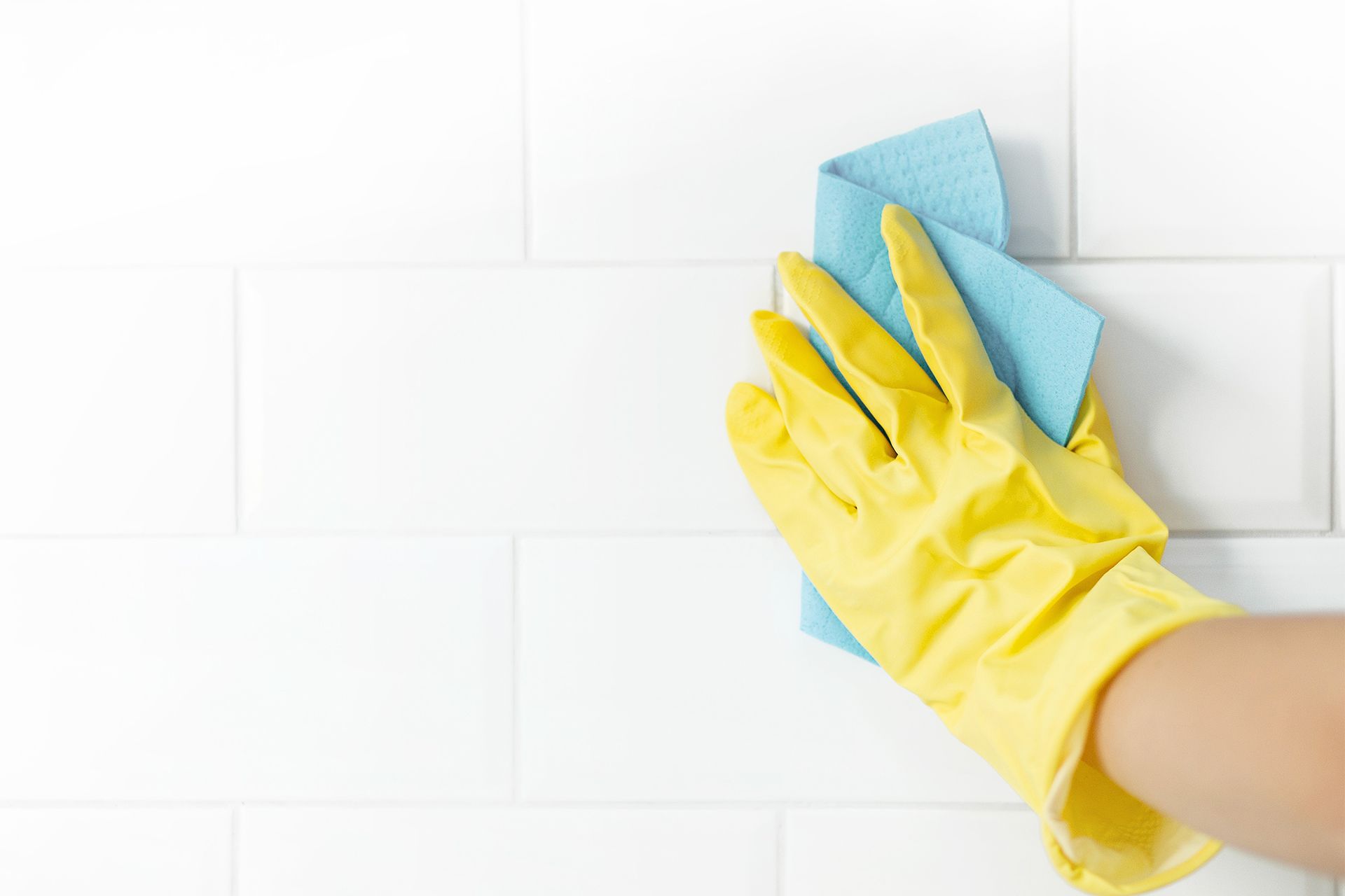 A person wearing yellow gloves is cleaning a white tile wall with a blue cloth.