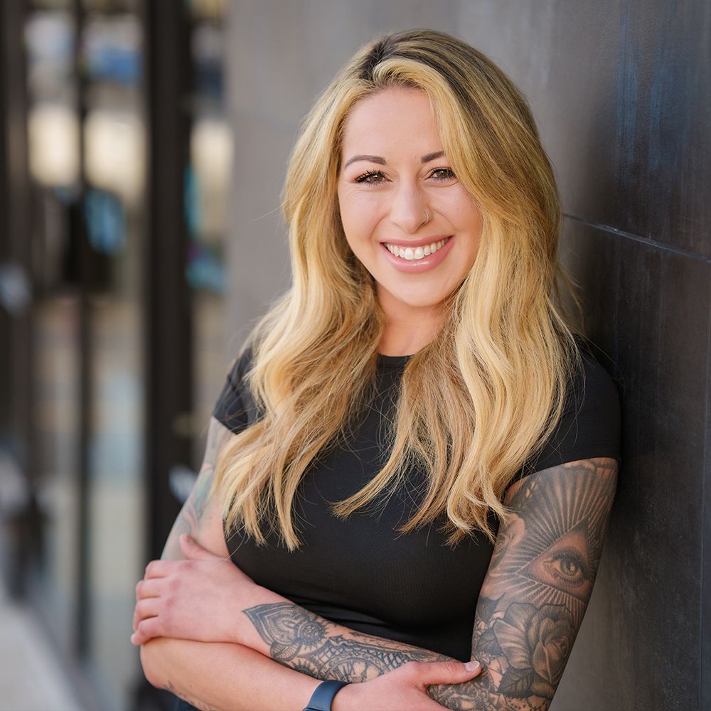 A woman with a tattoo on her arm is leaning against a wall and smiling.