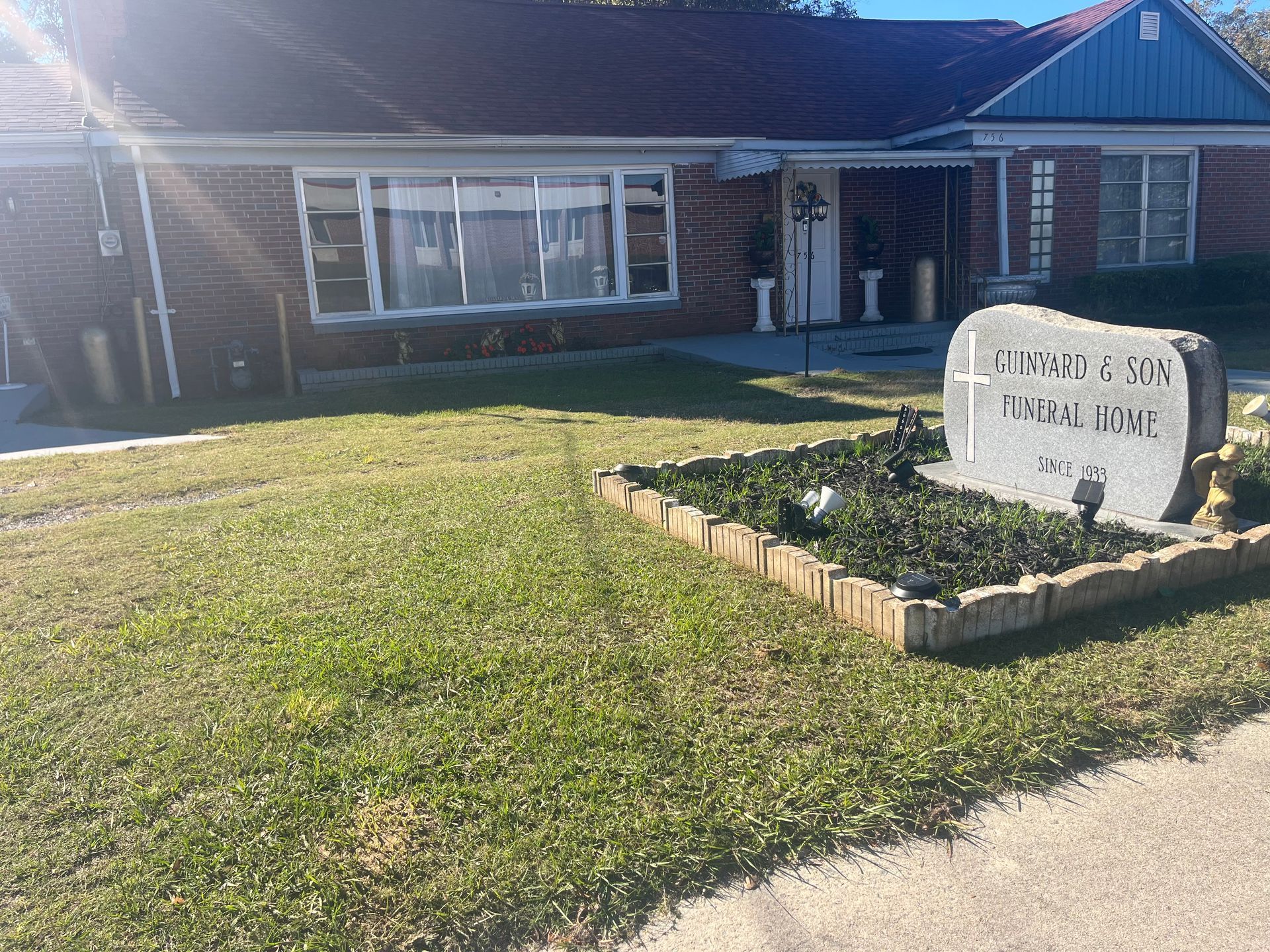 Exterior view of a brick building with a sign for a funeral home in front, with a headstone and green lawn.