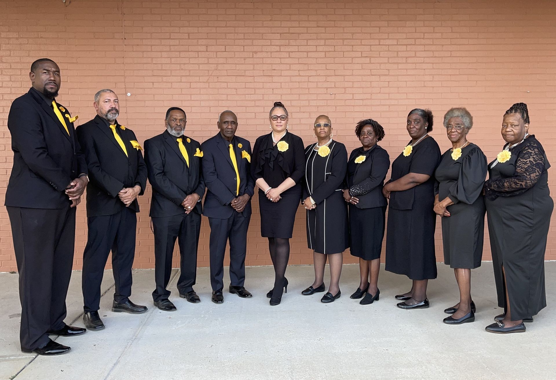 Group of people in black attire with yellow accents standing in front of a brick wall.