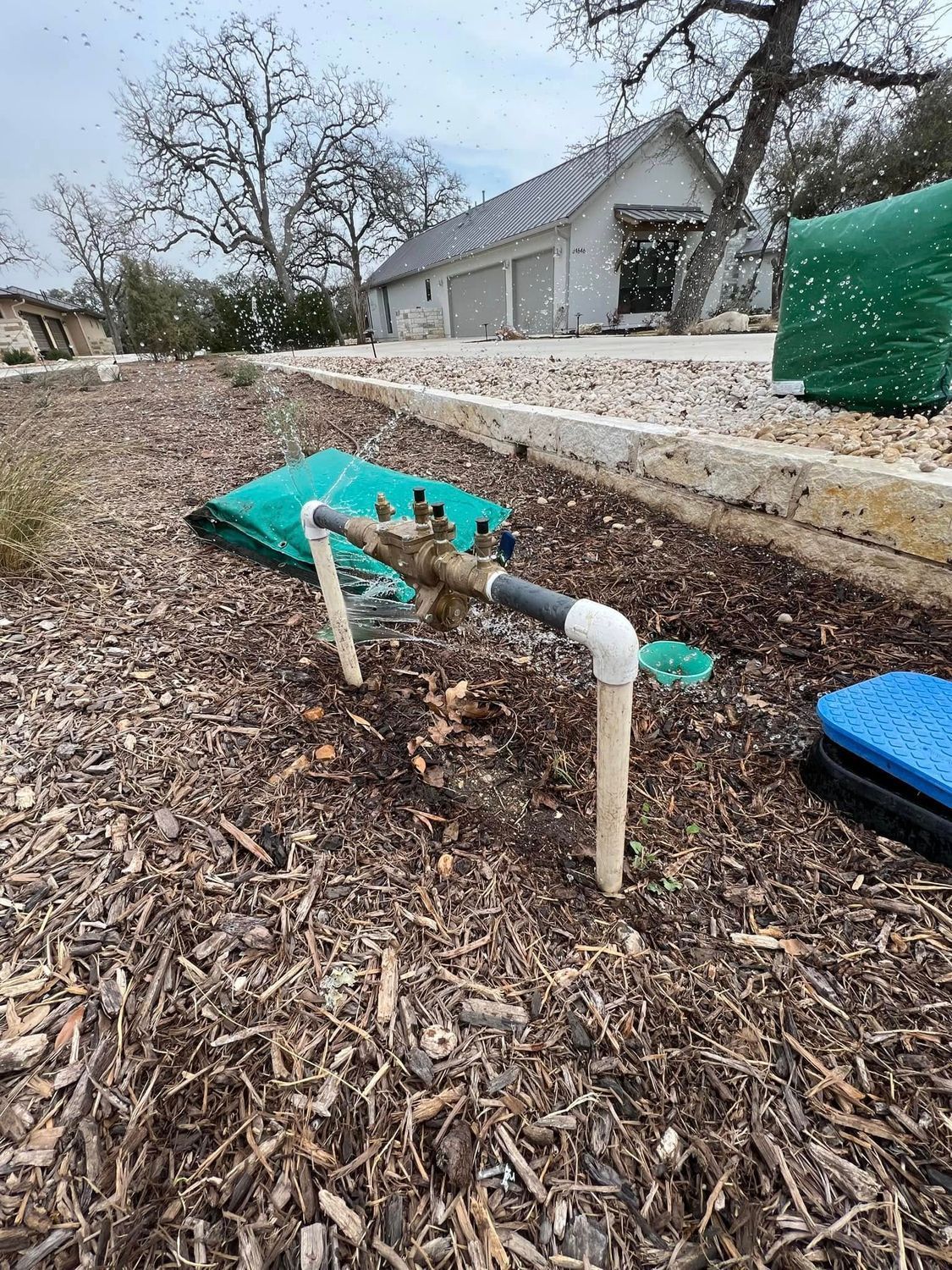 Irrigation system with exposed pipes and valves in a mulch bed in front of a house with a white garage.