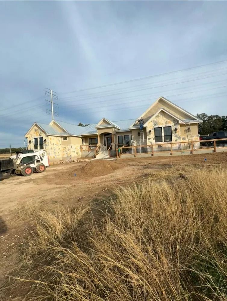 House under construction on a dirt lot, with a skid steer loader. Dry grass in foreground.