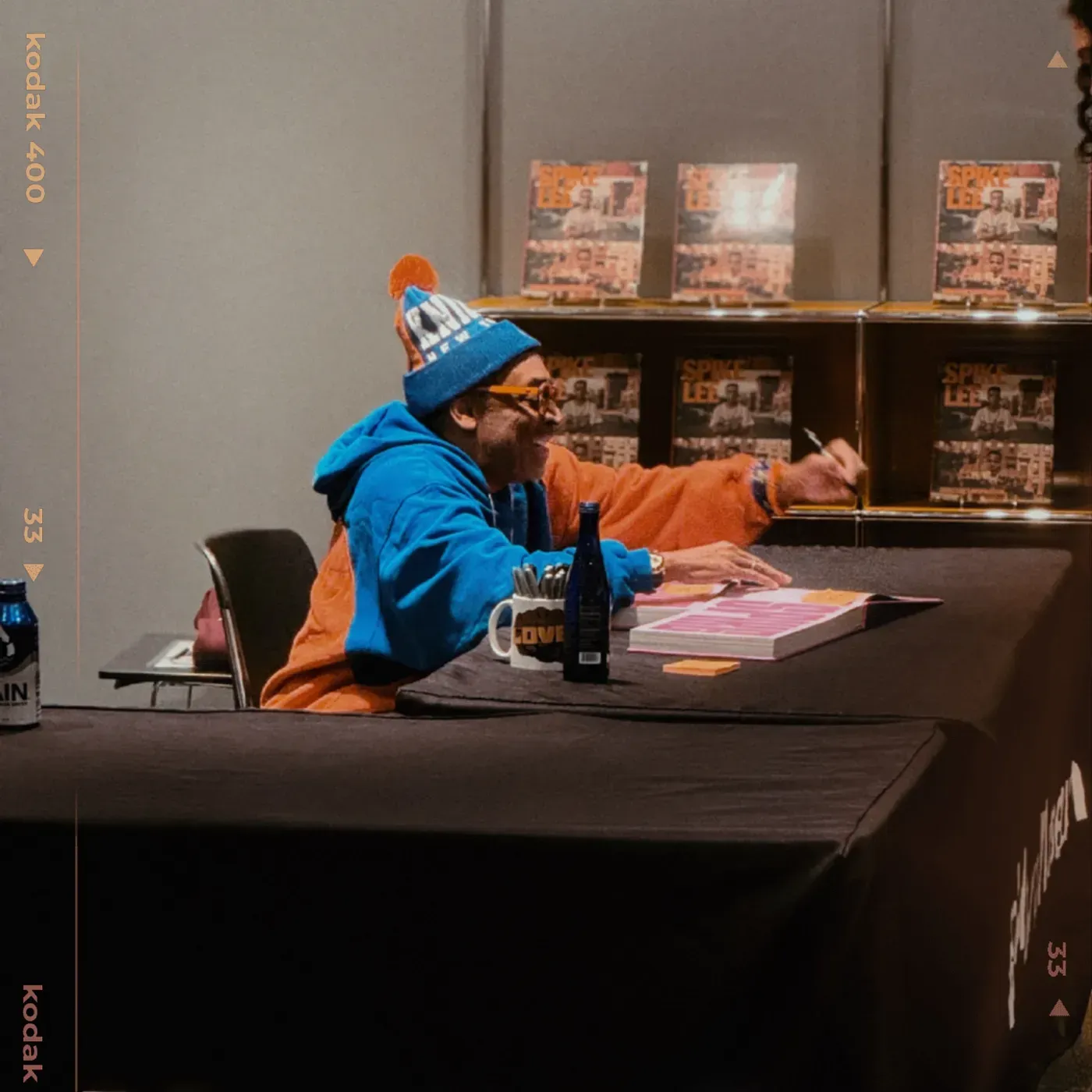 Person in a blue and orange outfit signing a book at a table. Shelves with books are in the background.