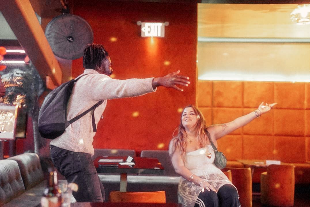 A man with a backpack is standing next to a woman sitting at a table in a restaurant.