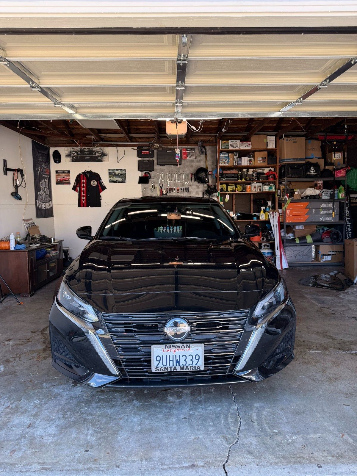 A black Nissan sedan parked in a garage cluttered with shelves, tools, and wall decorations.