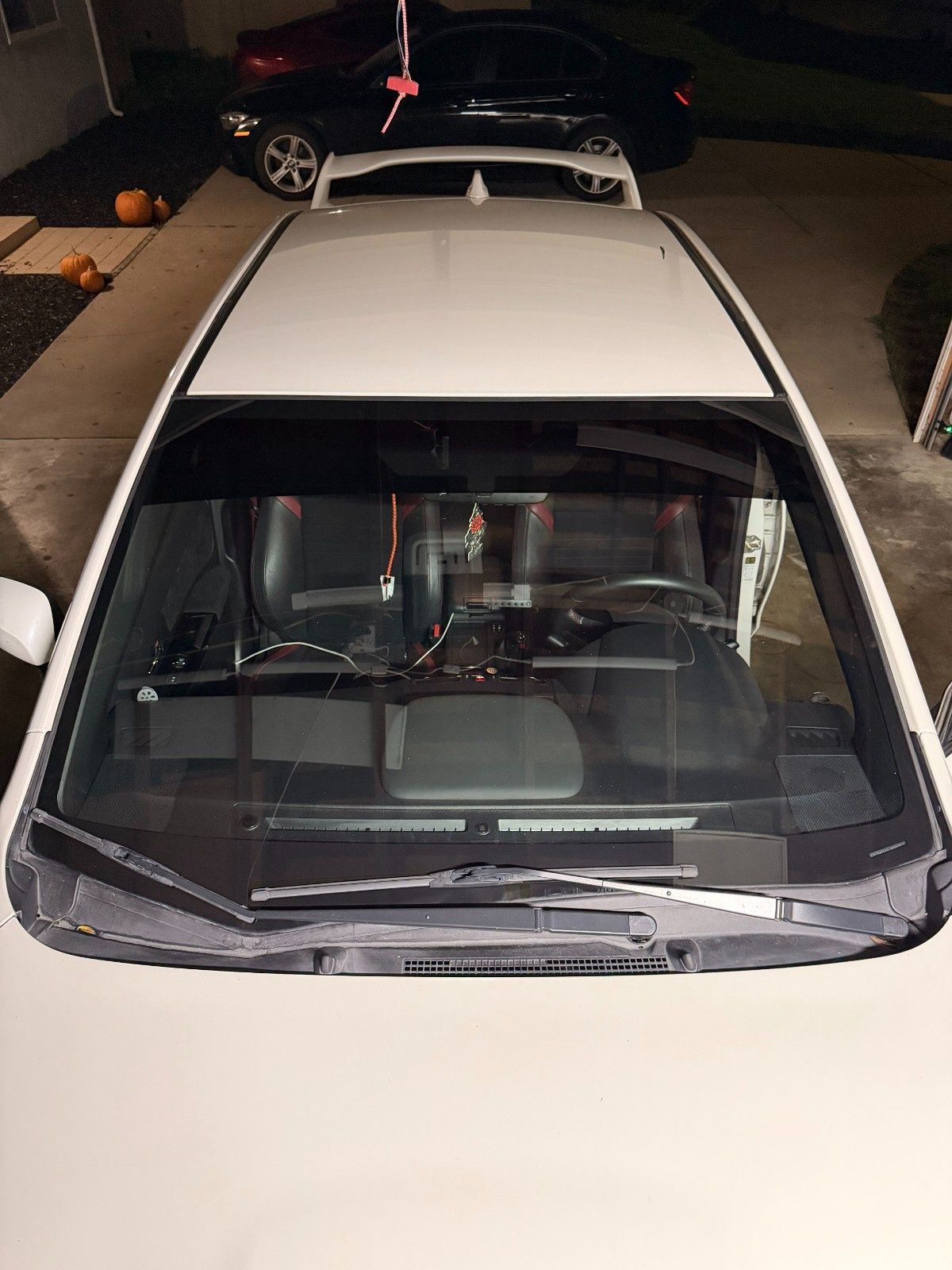 High-angle view of a white car’s windshield and roof, with a spoiler and a black car parked behind it in a driveway.