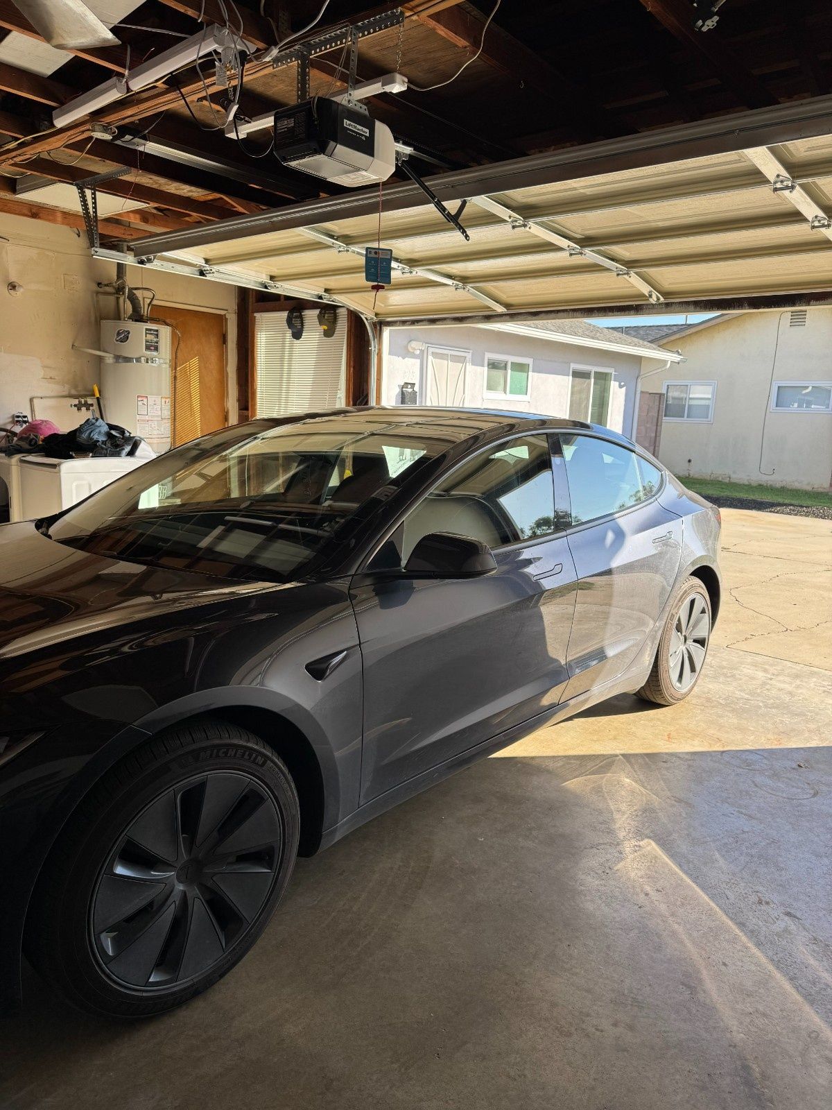 A dark grey Tesla parked inside a garage with the overhead door open, showing a driveway and house exterior beyond.