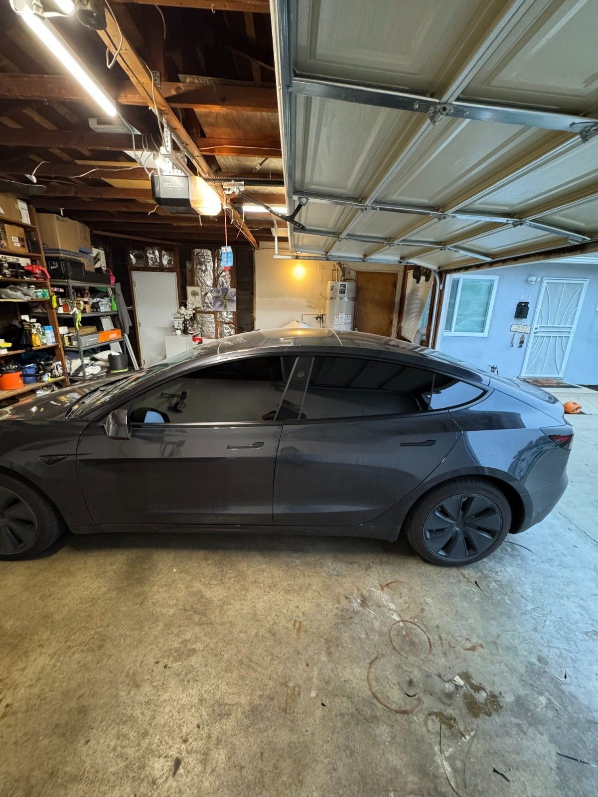 A dark gray Tesla Model 3 parked inside a residential garage with open shelving and a visible house exterior.