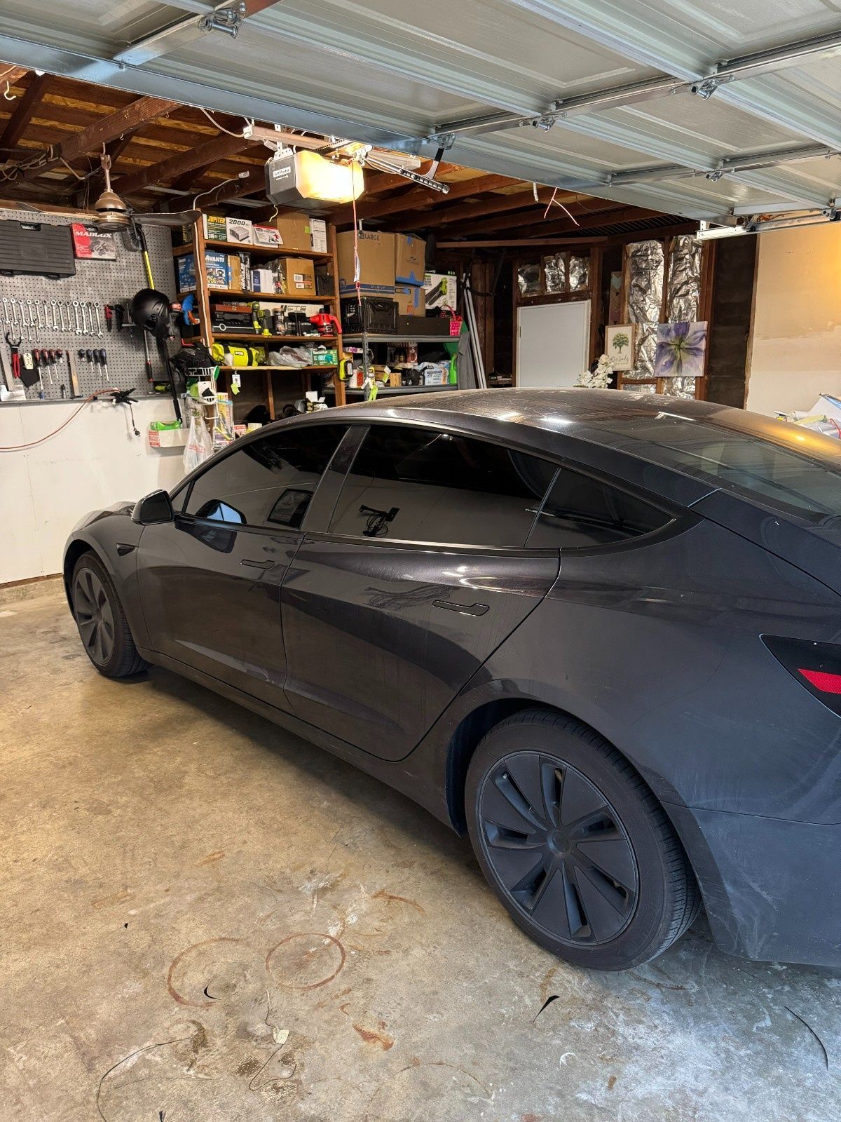 A dark gray Tesla Model 3 parked in a residential garage with storage shelving in the background.