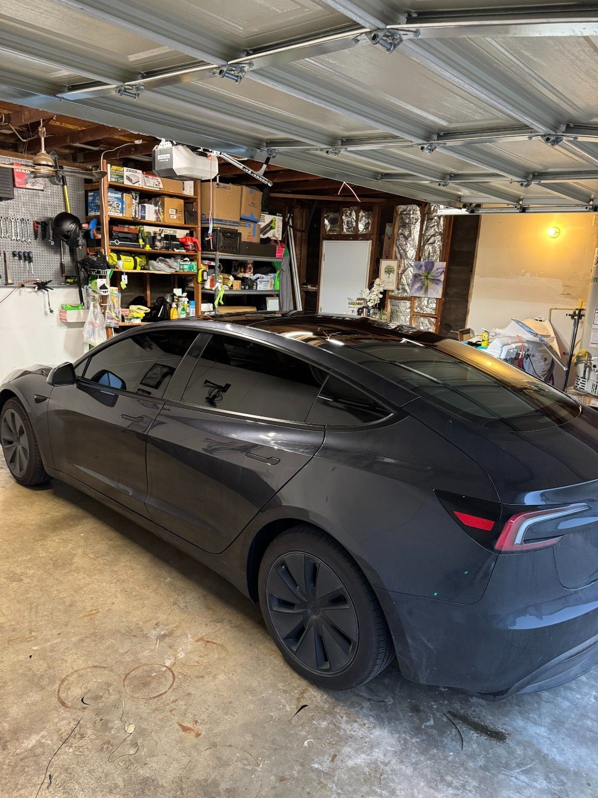 A dark gray Tesla parked inside a garage filled with storage shelves and various items.