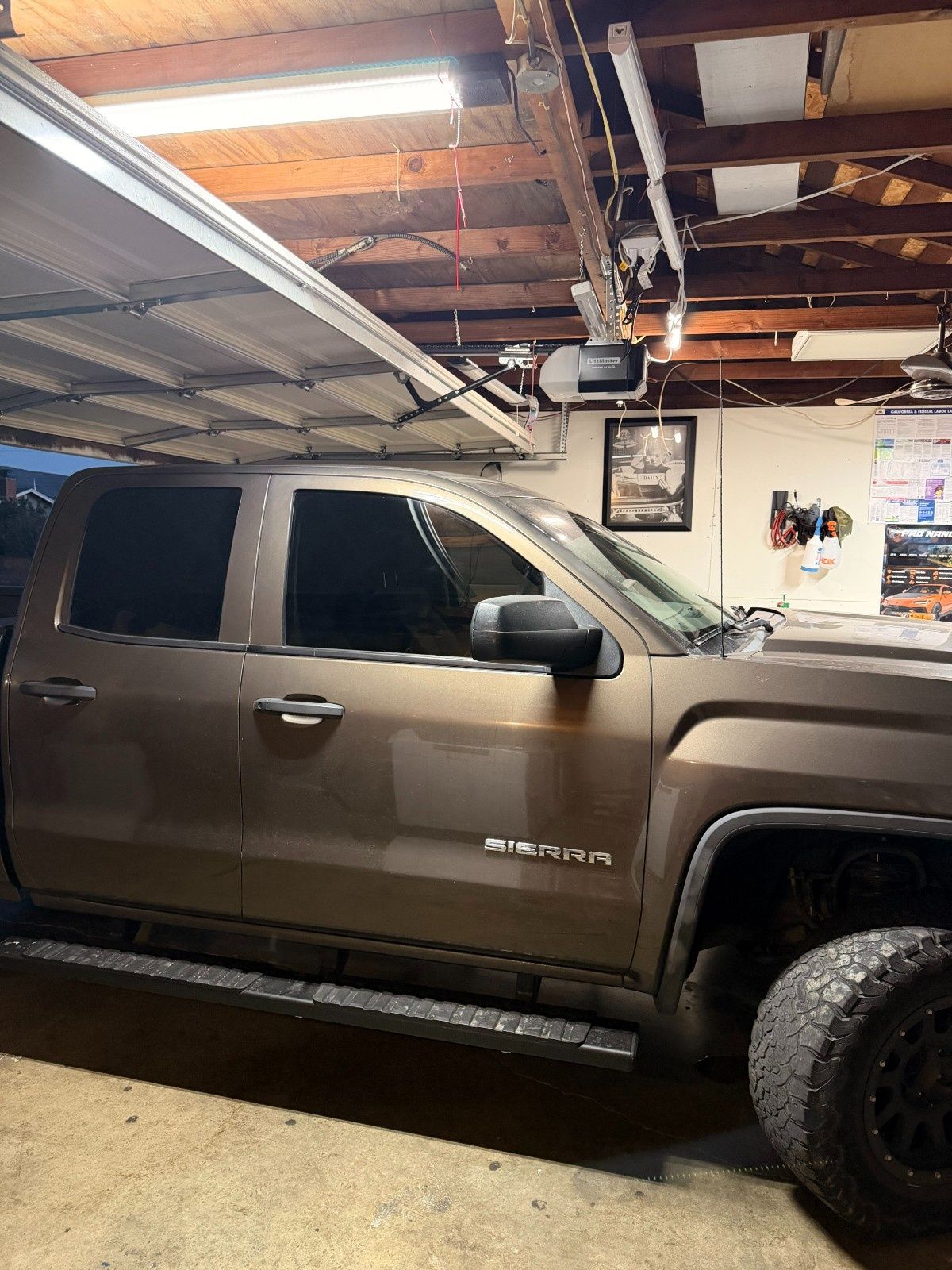 A brown pickup truck parked in a garage with wooden ceiling beams, a hanging light fixture, and an open garage door.