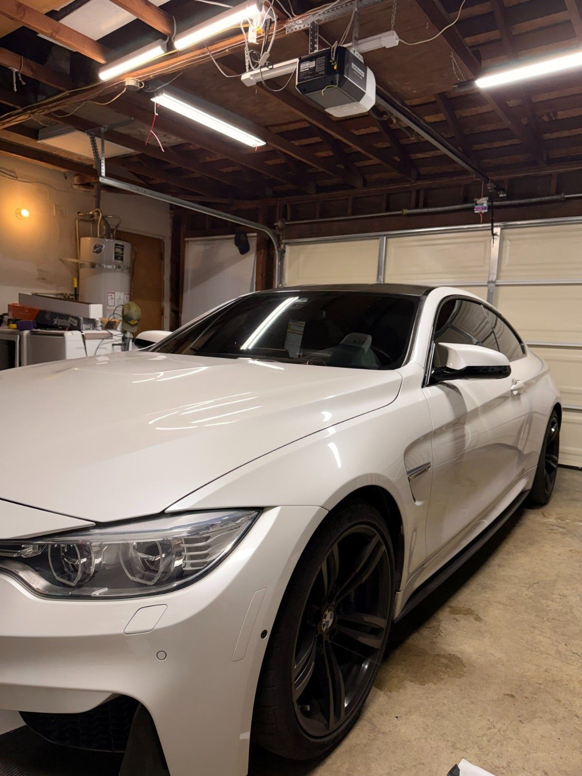 A white BMW M4 coupe with black wheels parked inside a residential garage with exposed wooden beams and LED lighting.