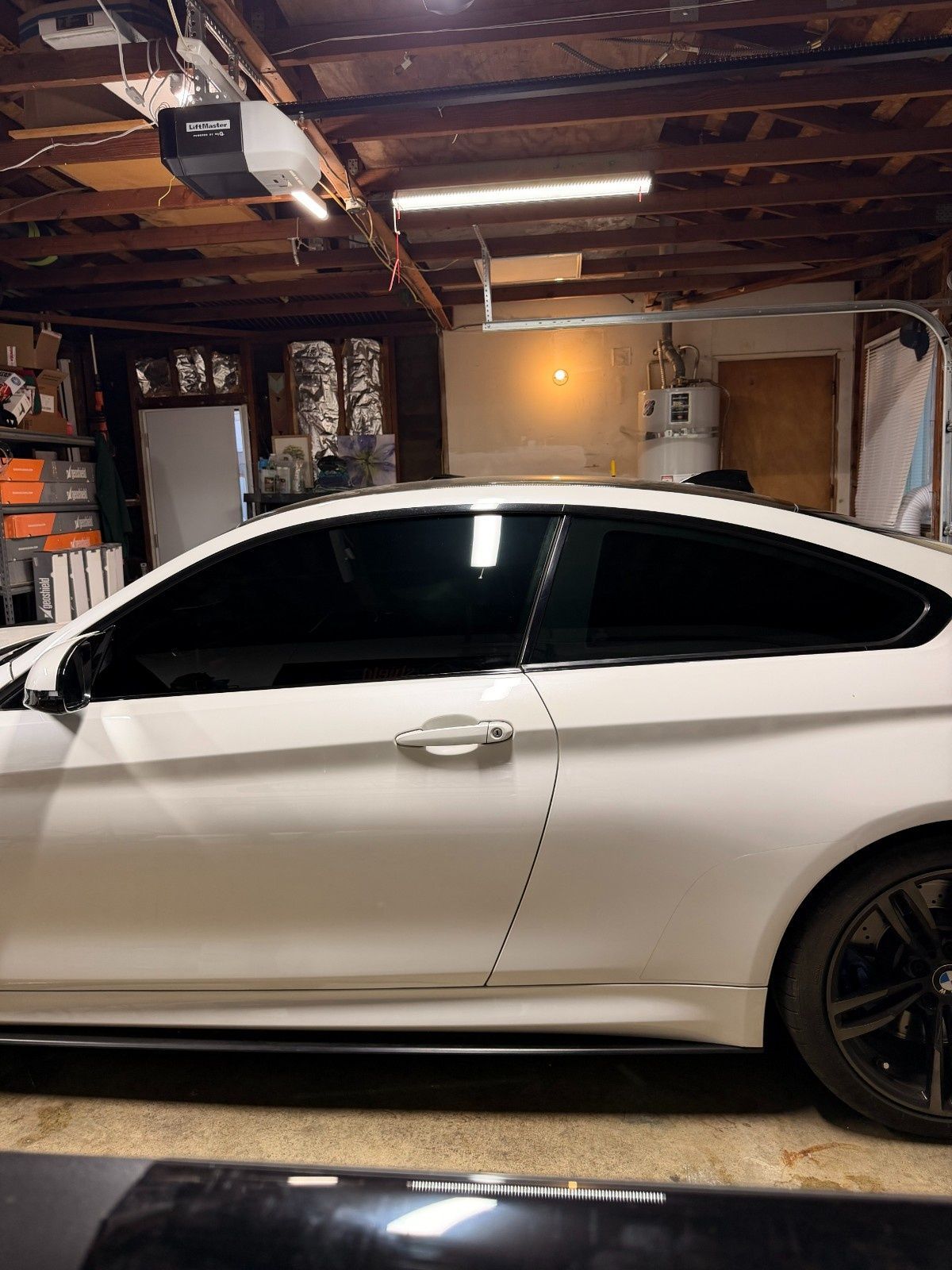 A white two-door coupe parked inside a dimly lit residential garage with exposed ceiling beams and concrete floors.