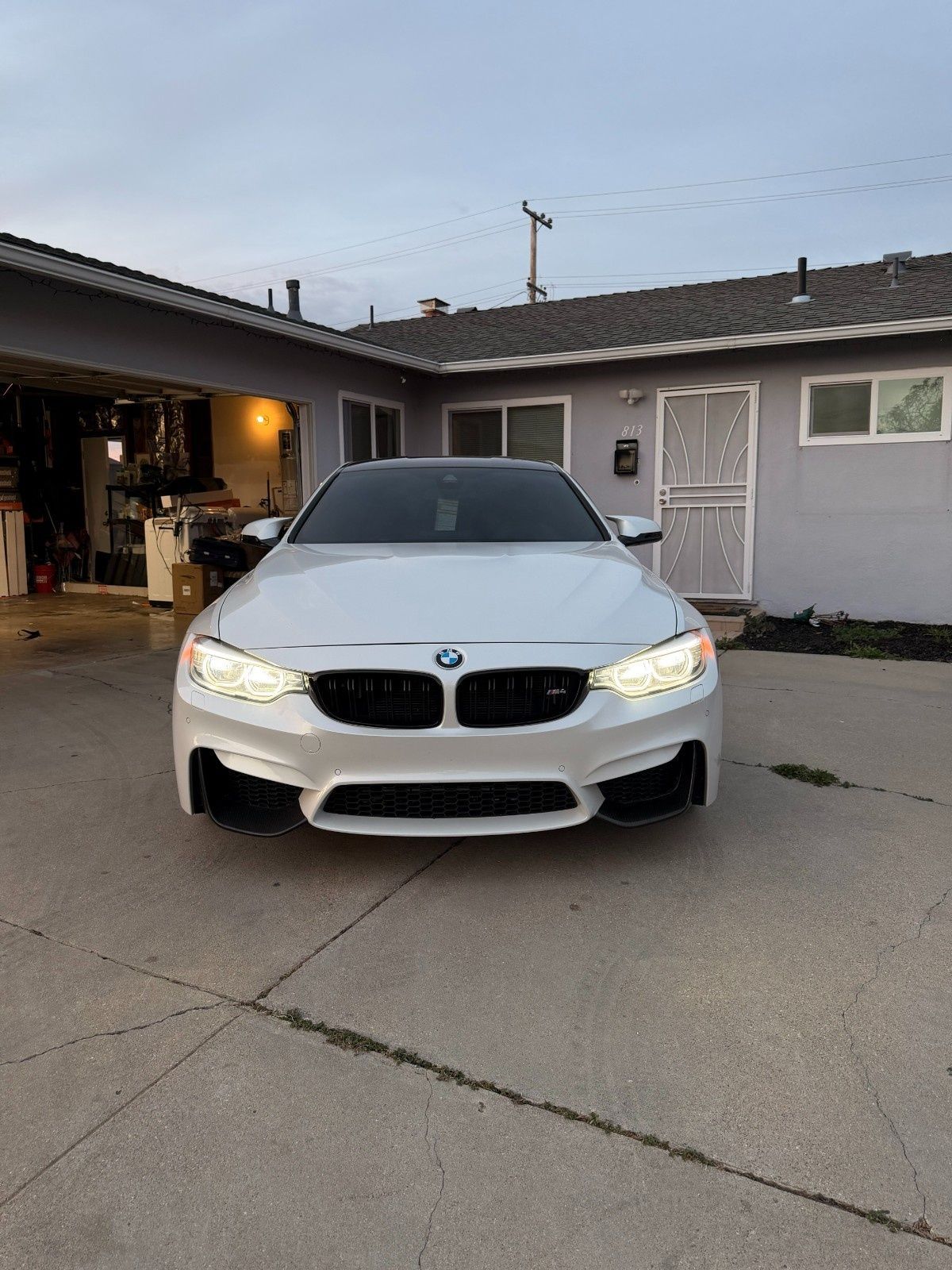 A white BMW M3 parked on a concrete driveway in front of a house at dusk.