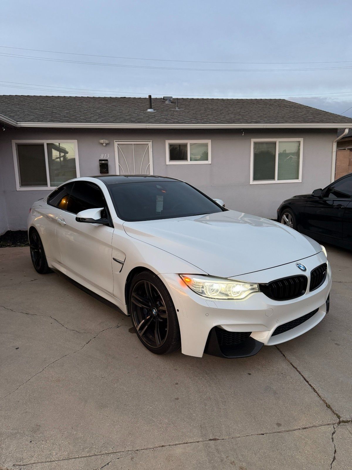 A white BMW M4 coupe parked on a concrete driveway in front of a single-story house.