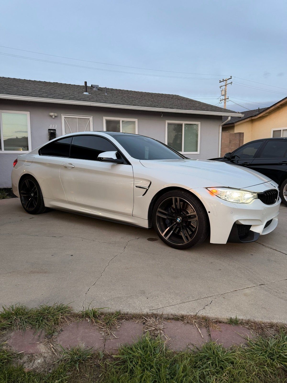 A white BMW M4 coupe parked on a concrete driveway in front of a gray house.