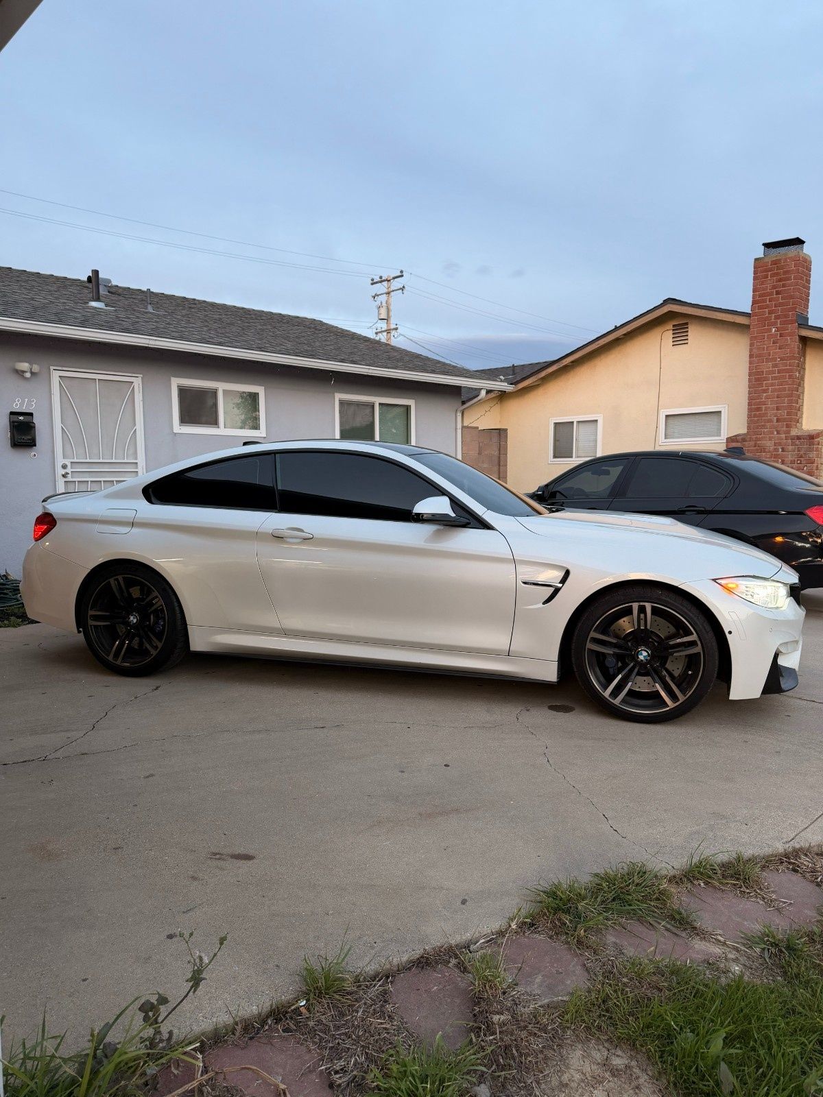 A white BMW M4 coupe parked on a concrete driveway in front of a house, with a black car partially visible behind it.