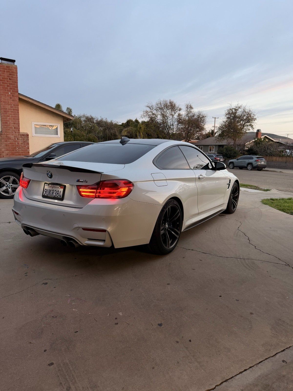 A white BMW M4 coupe parked on a residential driveway at dusk, featuring dark wheels and tinted windows.