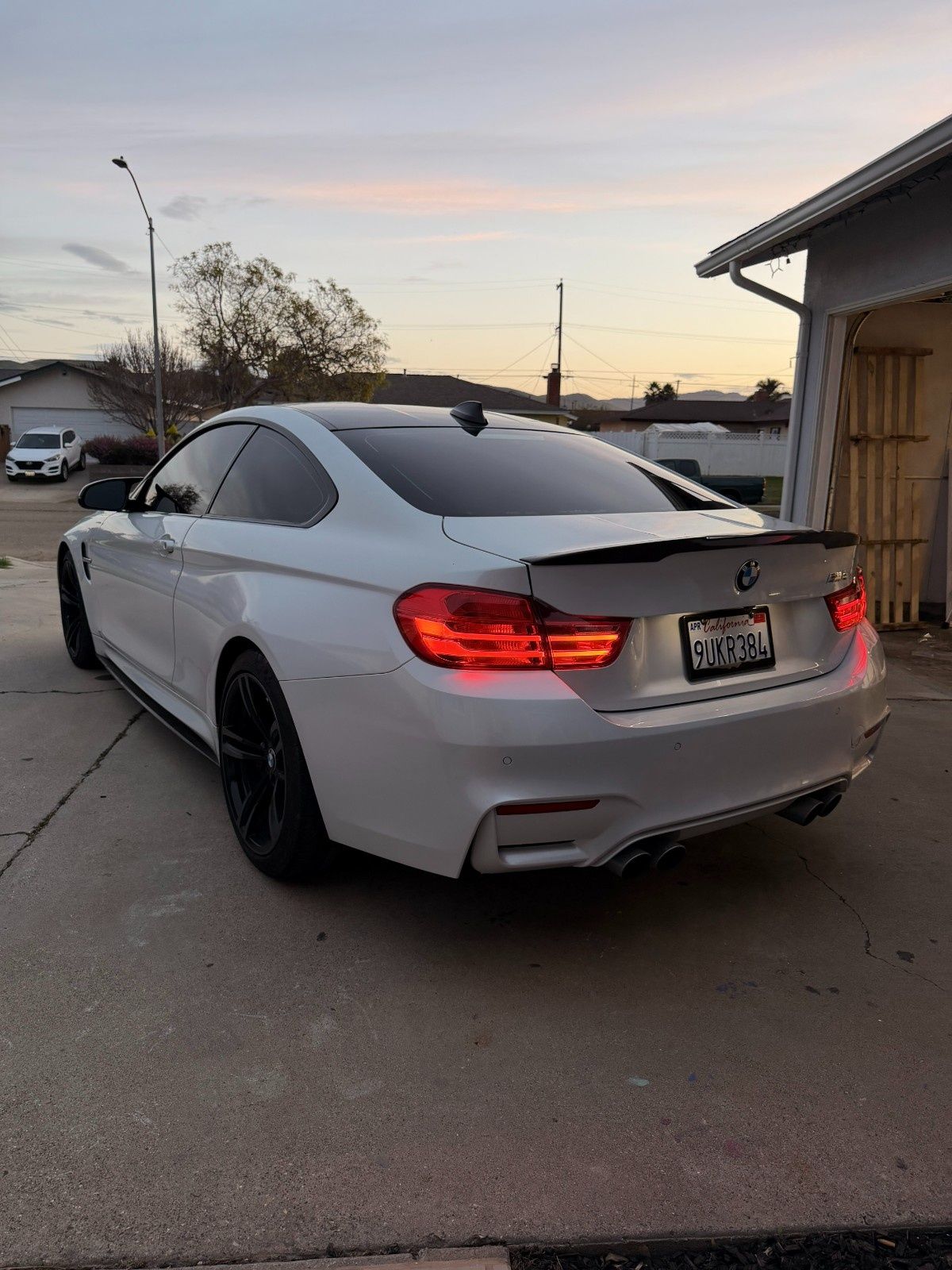 A white BMW coupe with black wheels and a rear spoiler parked in a driveway at sunset.