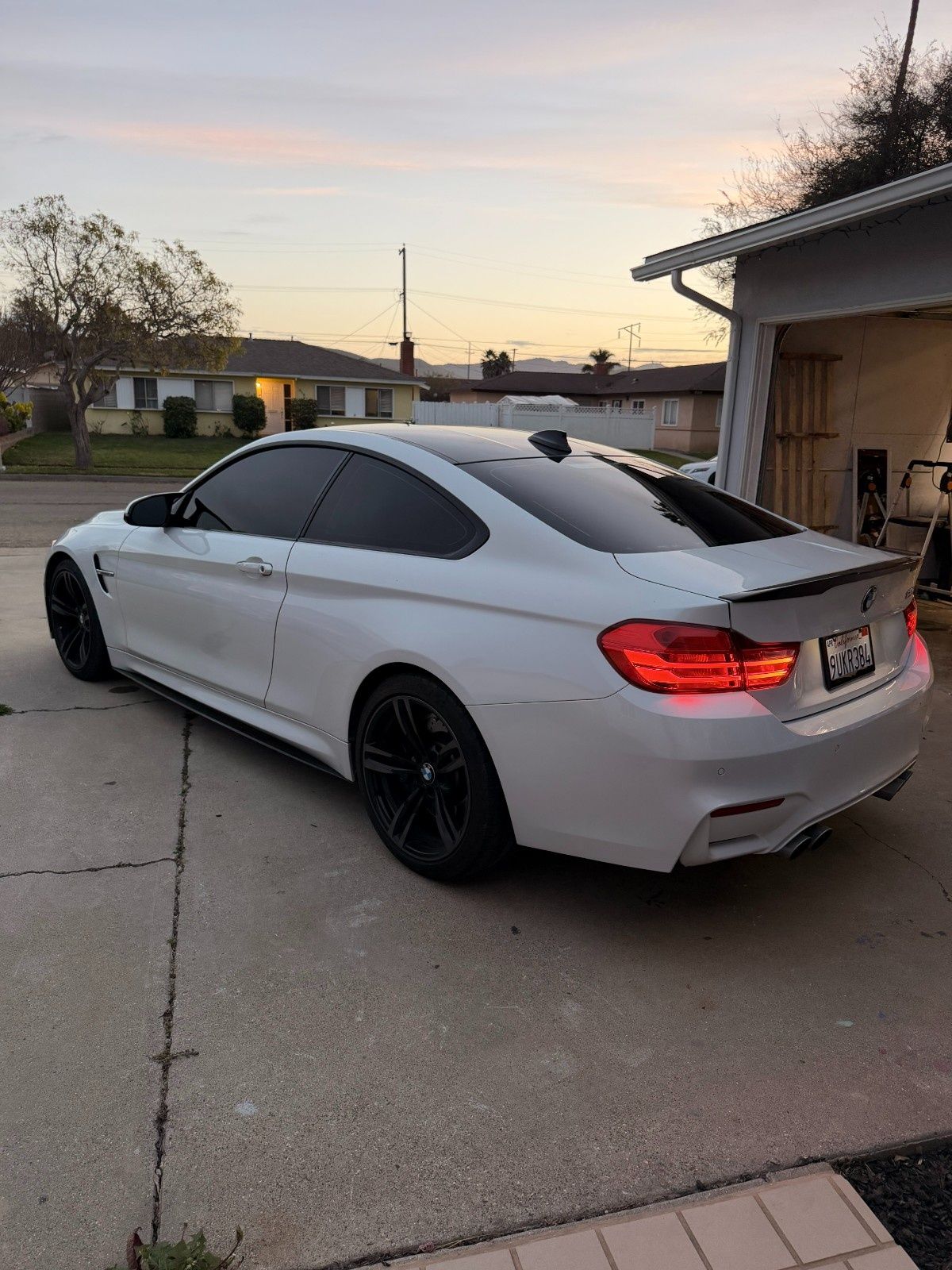 A white BMW coupe with black wheels and tinted windows parked on a residential driveway at sunset.