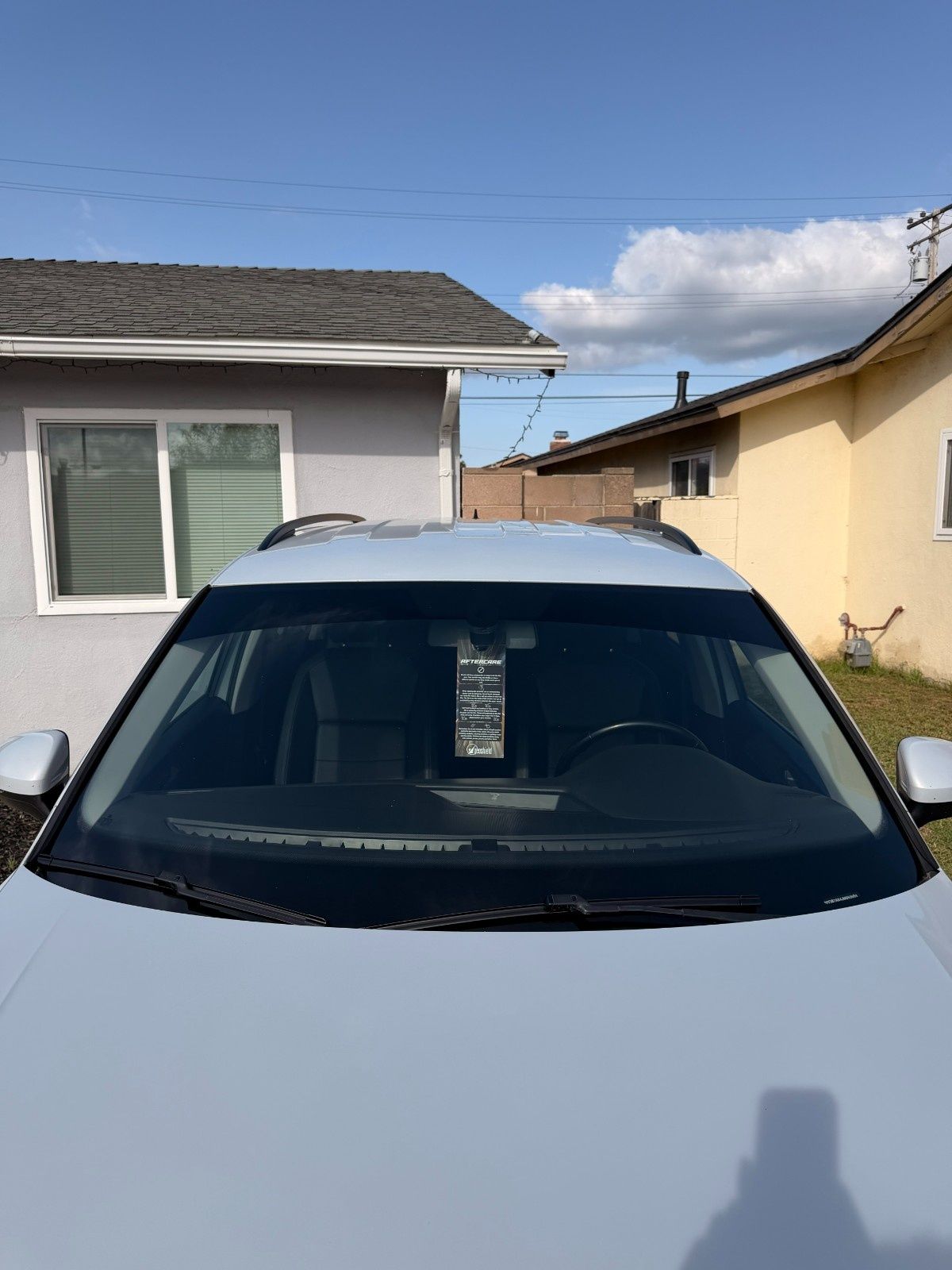 A silver car parked between two houses, viewed from the front, with a paper tag hanging from the rearview mirror.
