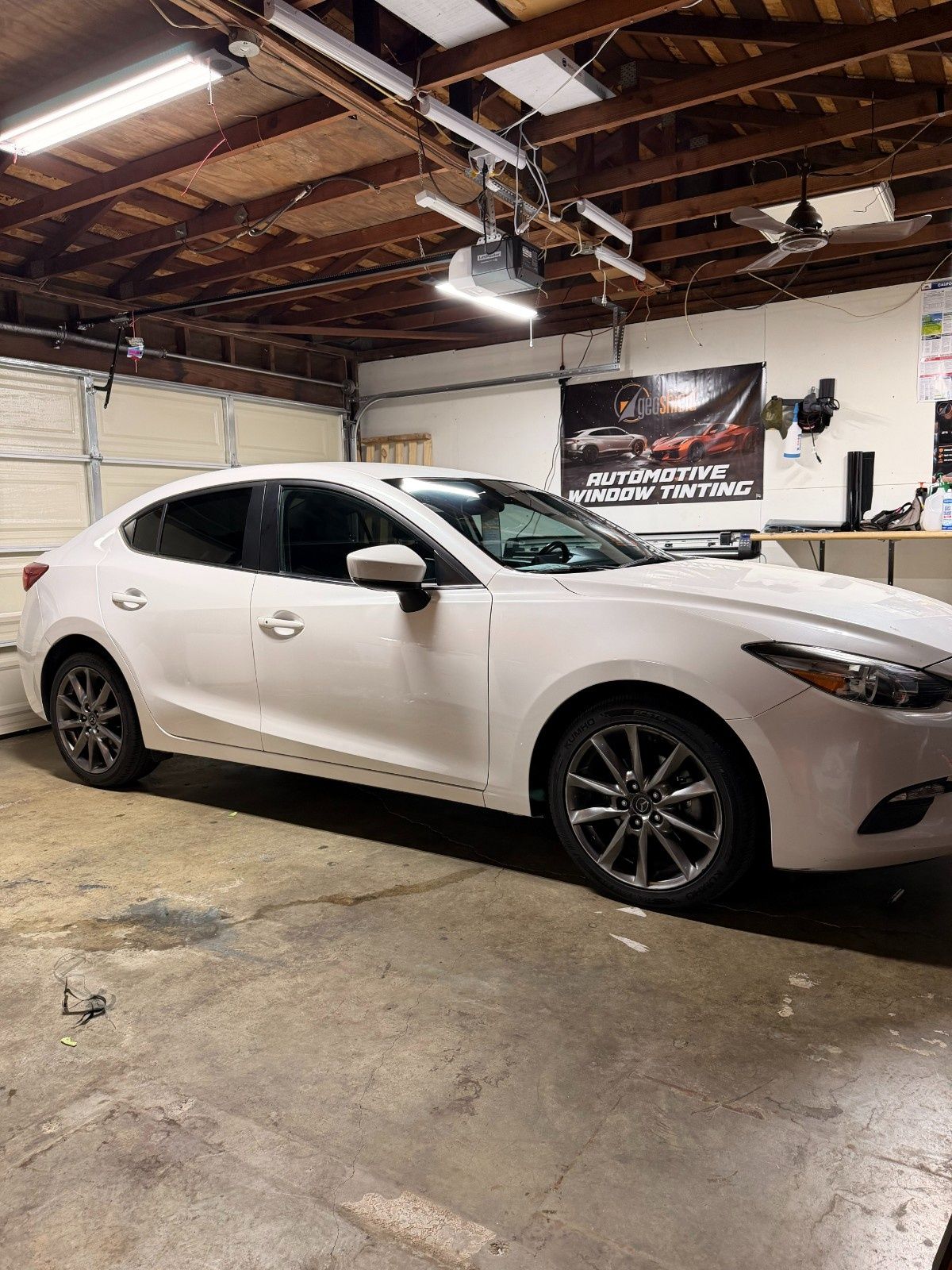 A white sedan parked inside a garage with workshop lights and a sign on the back wall.