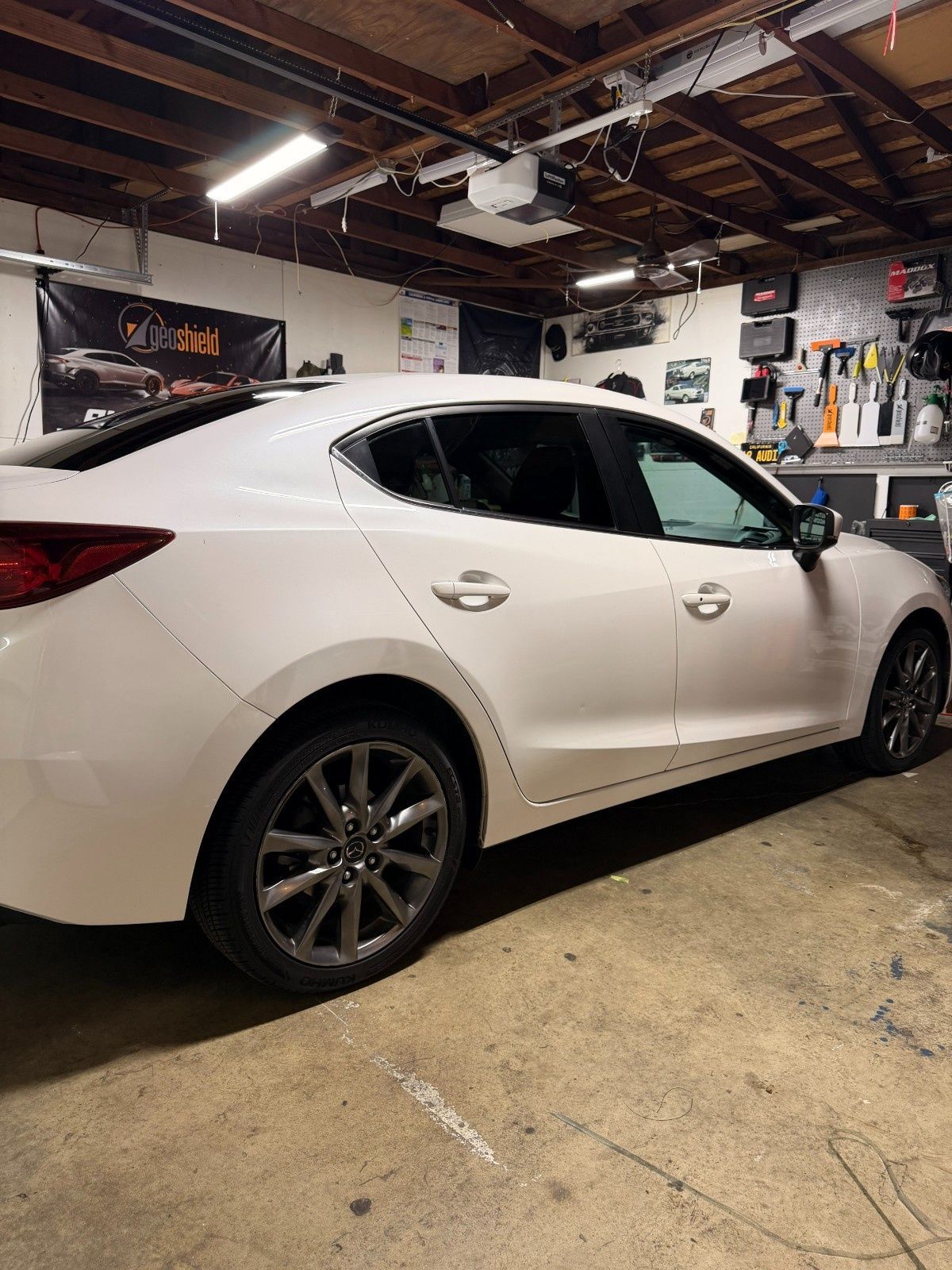 A white sedan parked inside a garage with workshop tools and lighting visible in the background.