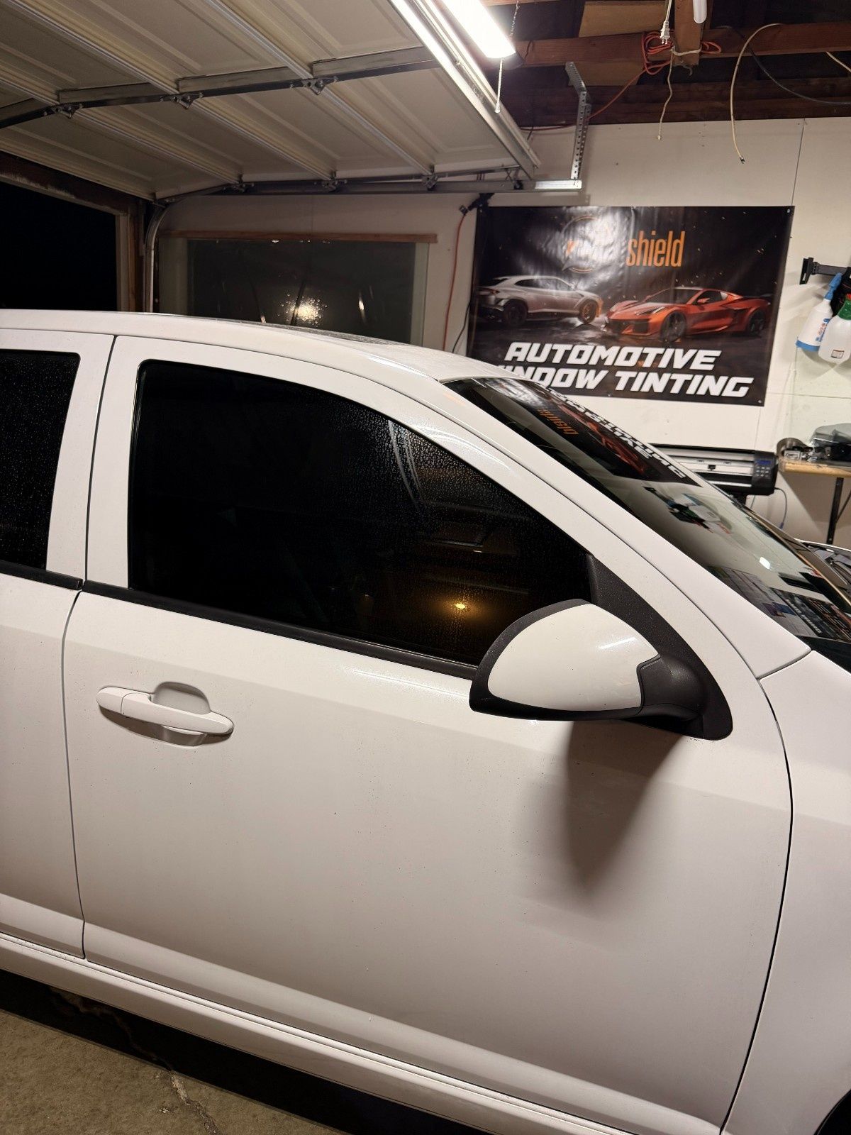 White car with tinted windows parked in a garage with an automotive window tinting sign in the background.