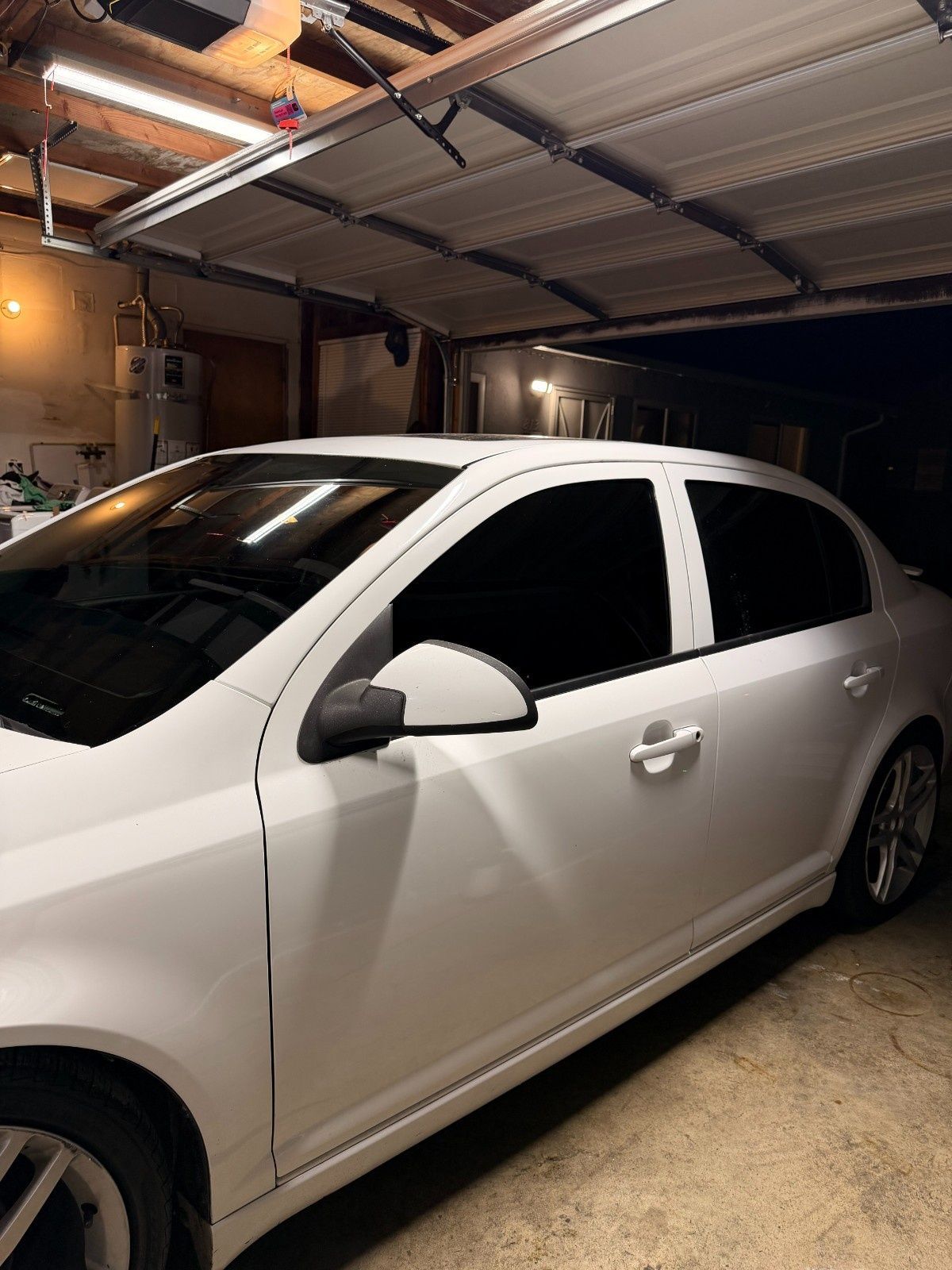 A white sedan parked inside a garage at night, illuminated by overhead lighting.
