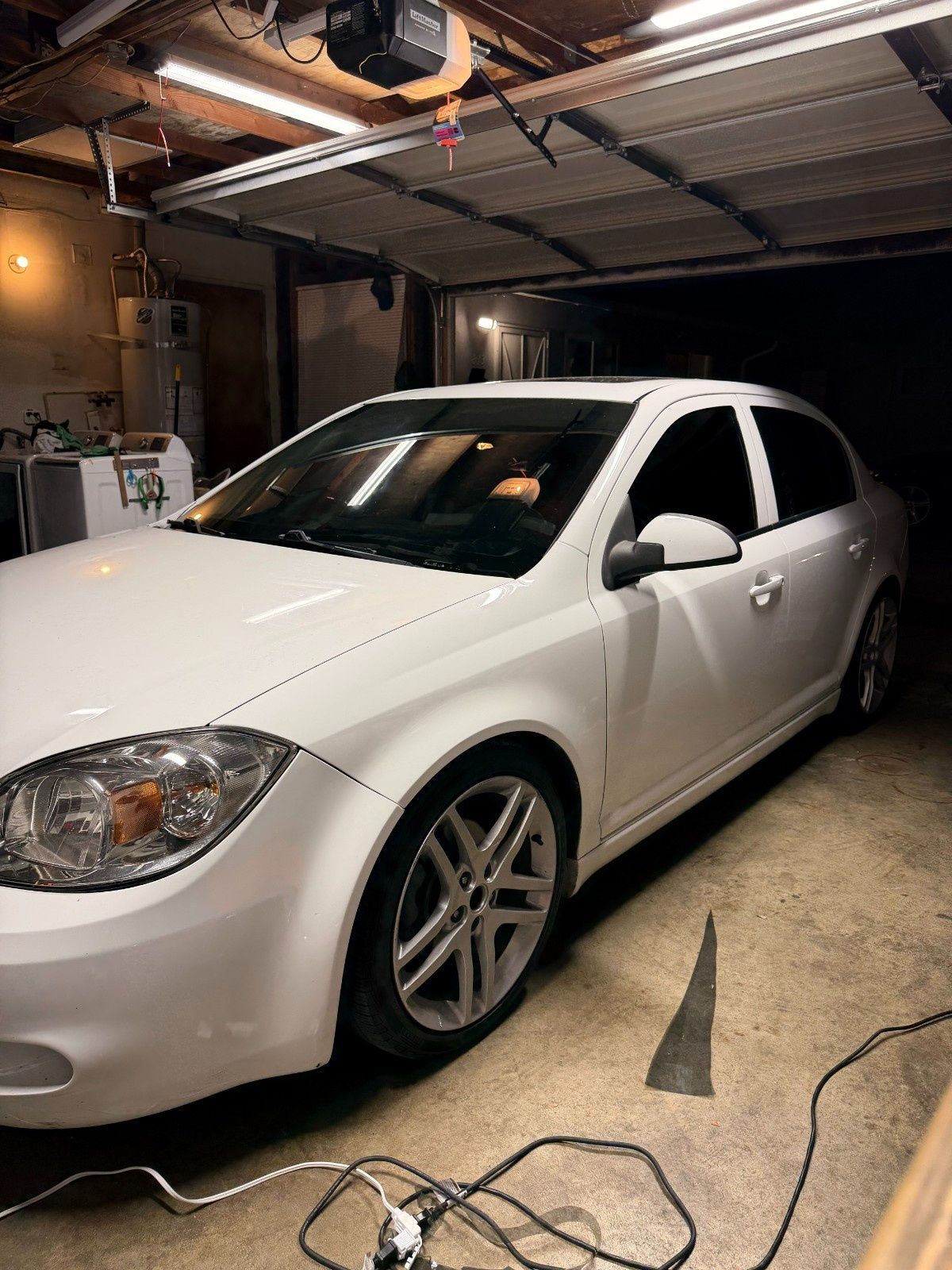 A white sedan parked inside a garage with its headlights reflecting overhead lights.