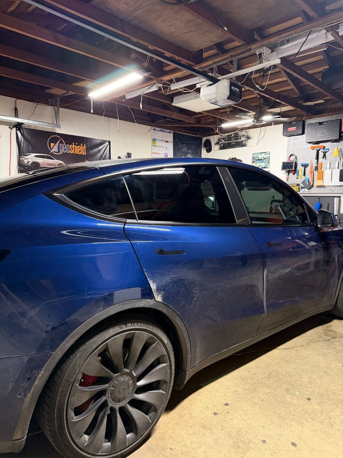 A blue Tesla parked in a cluttered garage, showing noticeable damage on the rear passenger door panel.