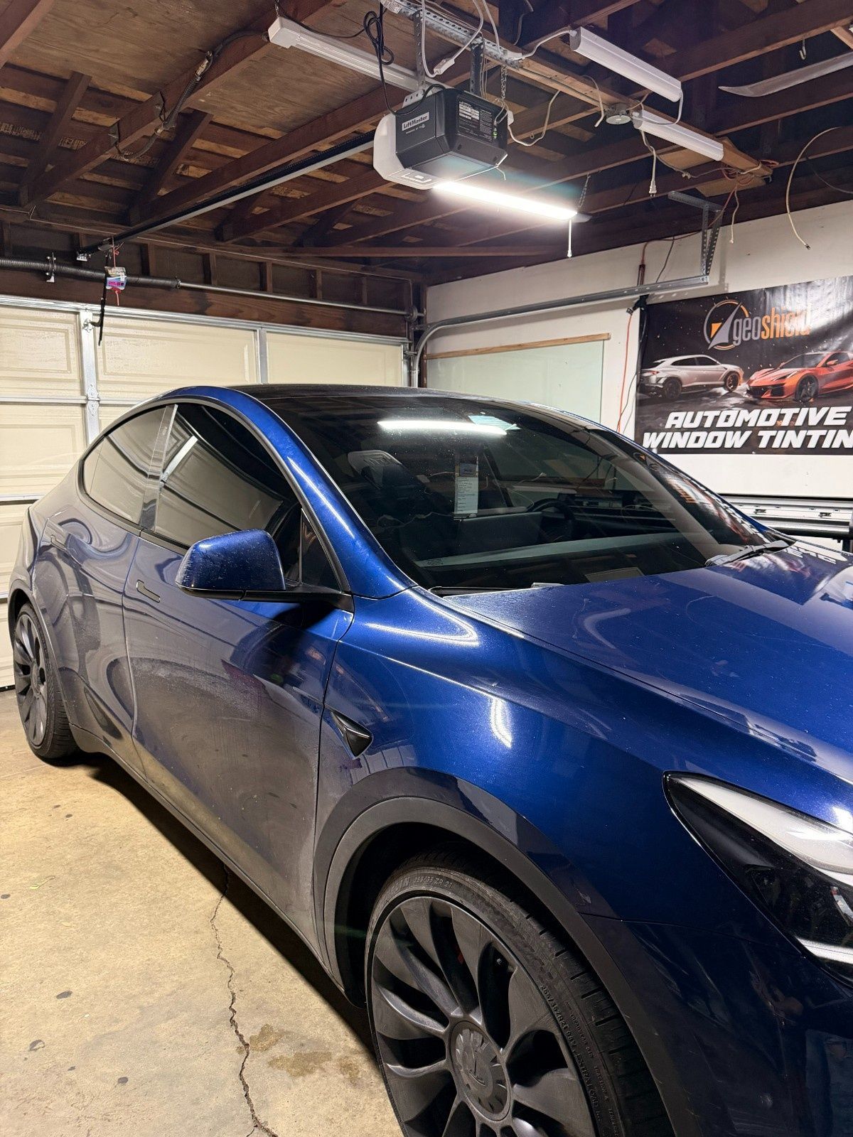 A deep blue Tesla parked in a garage, showcasing professionally tinted windows with a shop sign in the background.