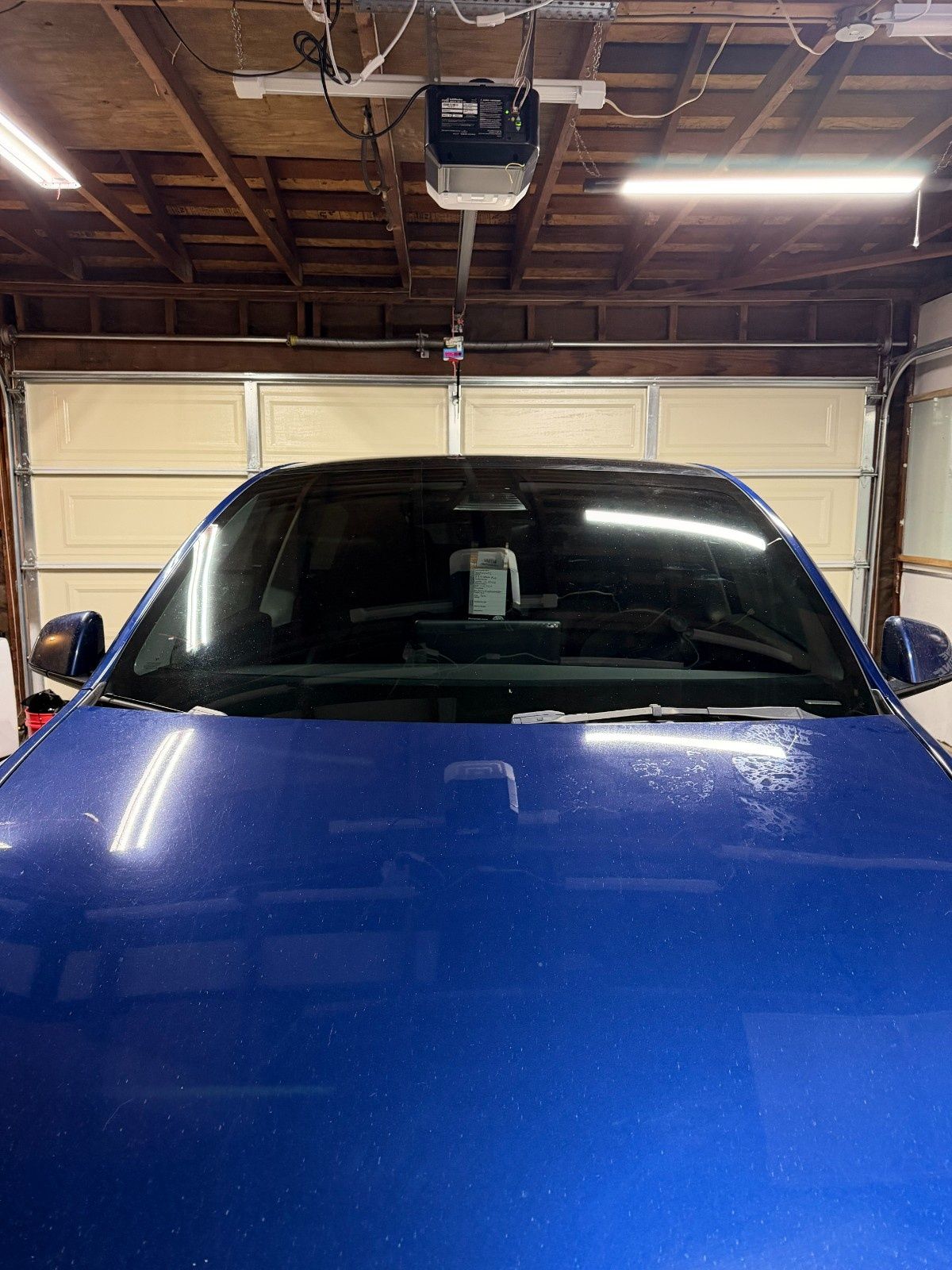 A blue car parked inside a garage, viewed from the front with a garage door opener hanging above.
