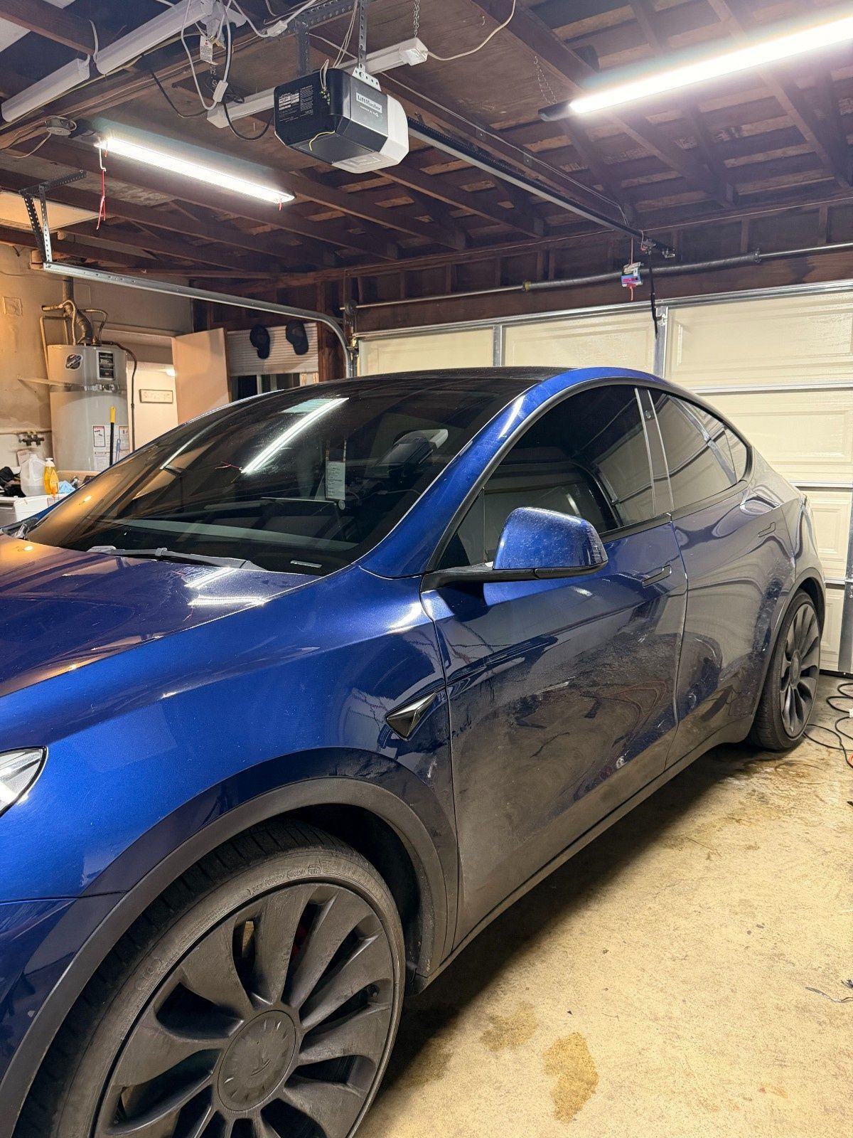 A dark blue Tesla parked inside a garage with an open door and fluorescent ceiling lights.