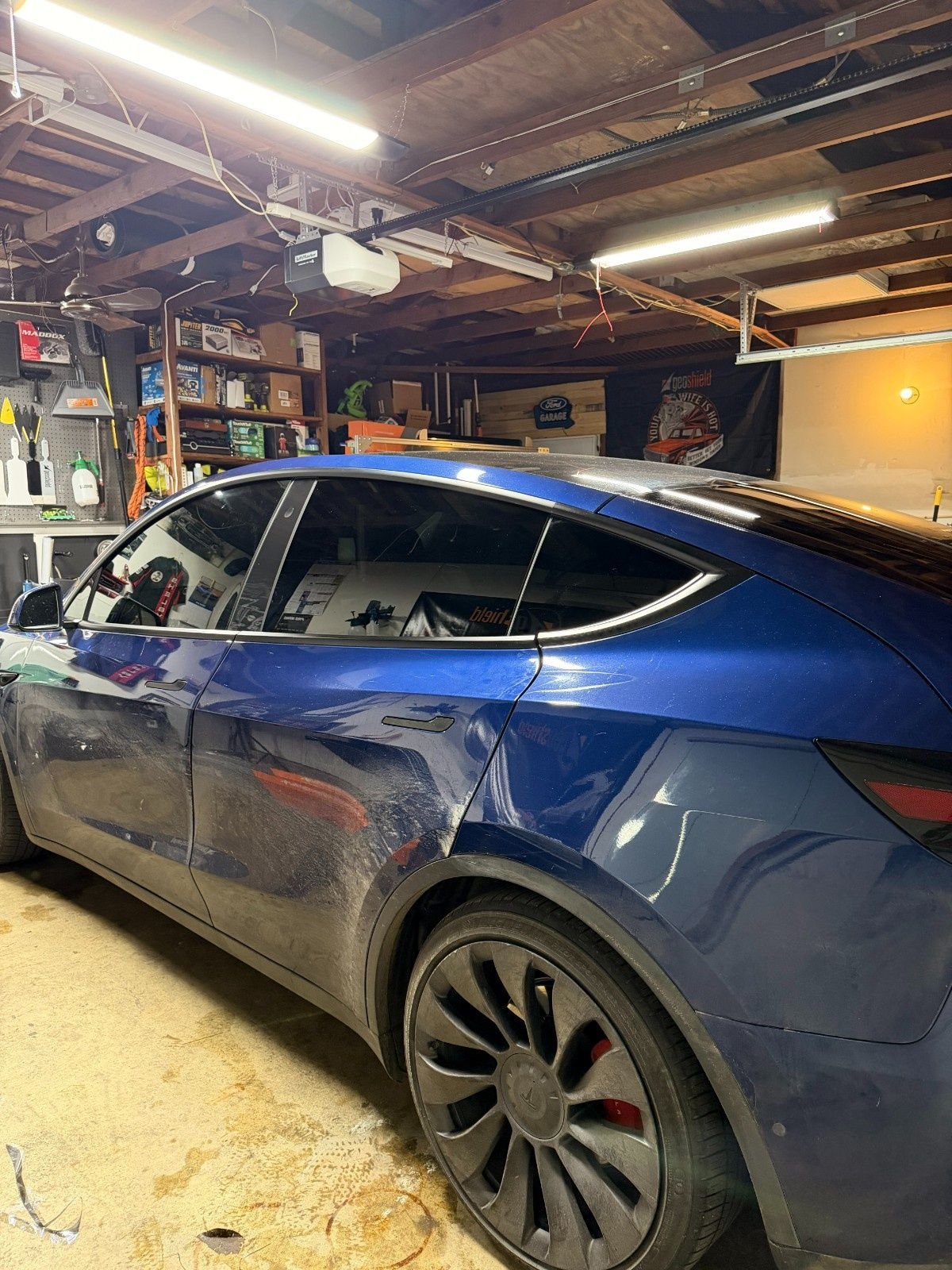 A deep blue Tesla parked in a cluttered garage, showing a significant dent on the rear passenger-side door and panel.