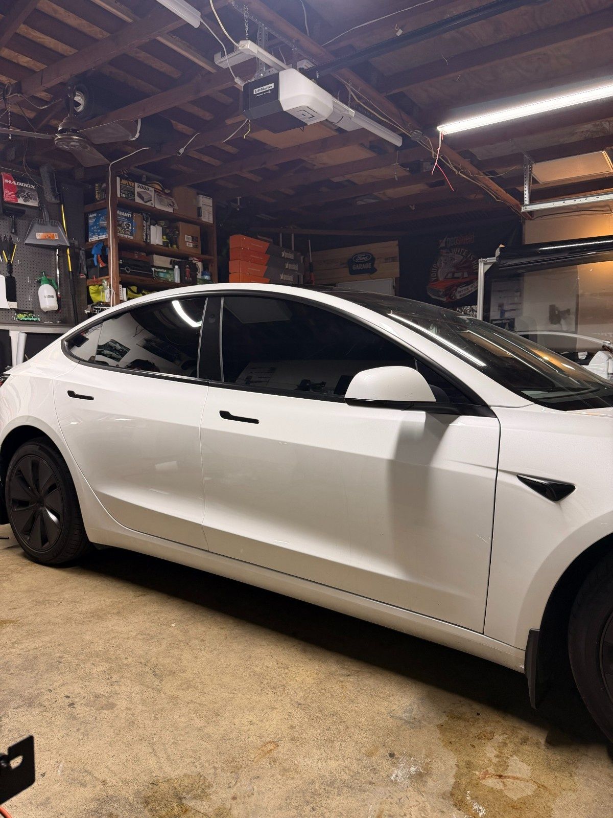 A white Tesla Model 3 with black wheels parked inside a cluttered residential garage.
