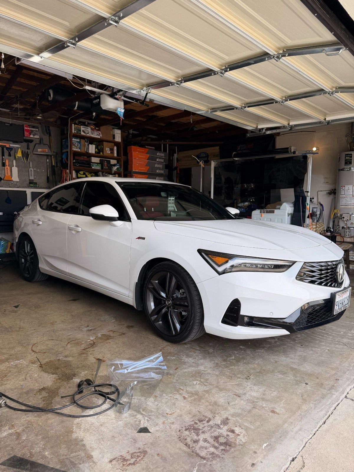 A white Acura Integra parked inside a residential garage with clutter on the shelves in the background.