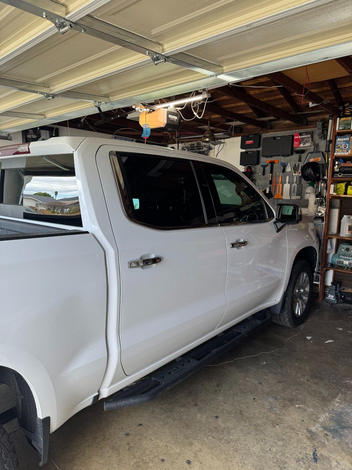 A side view of a white pickup truck parked in a garage, next to a shelving unit filled with tools and various items.