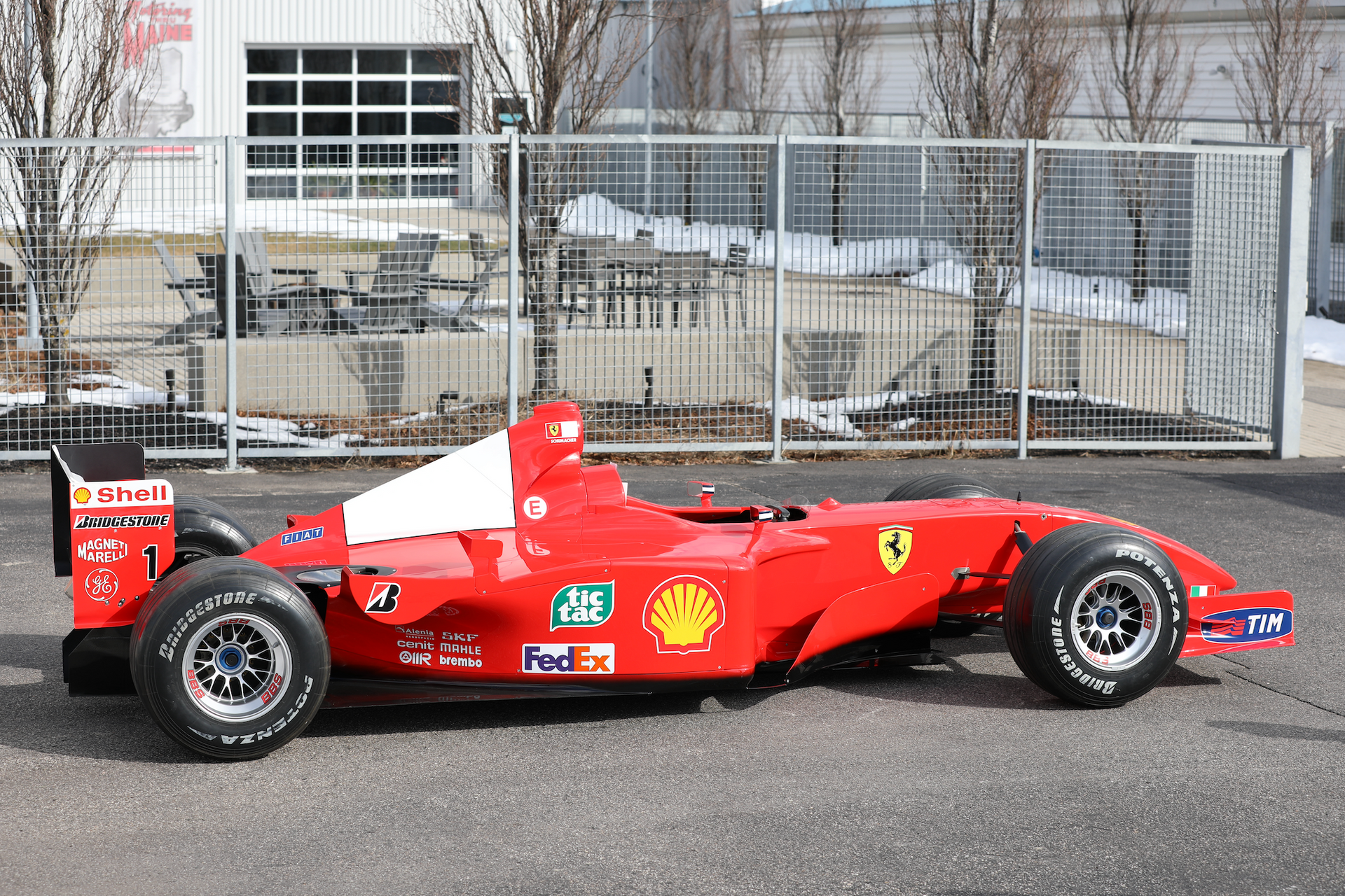 Red Formula 1 race car parked outdoors in front of a metal fence