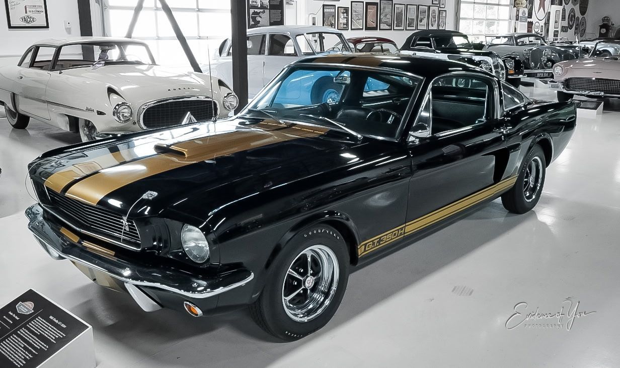 Black classic muscle car with gold racing stripes displayed indoors at an auto show