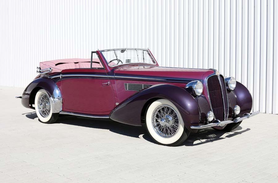 Vintage maroon convertible with whitewall tires parked outdoors against a white corrugated wall