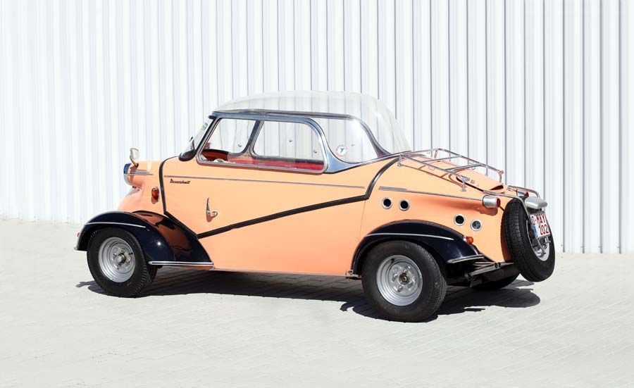 Vintage peach-and-black three-wheeled car parked on a light floor against a white corrugated wall.