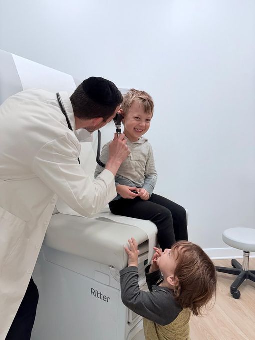 Doctor examining a smiling child’s ear while another child looks on in a clinic room