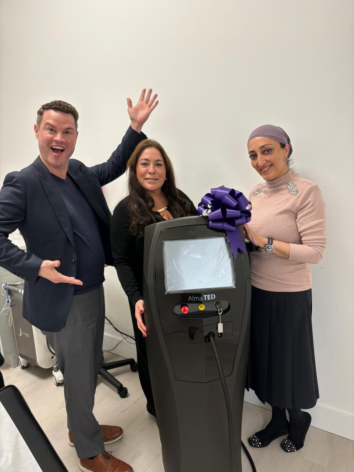 Three people pose beside a black machine with a purple ribbon bow in a plain room.