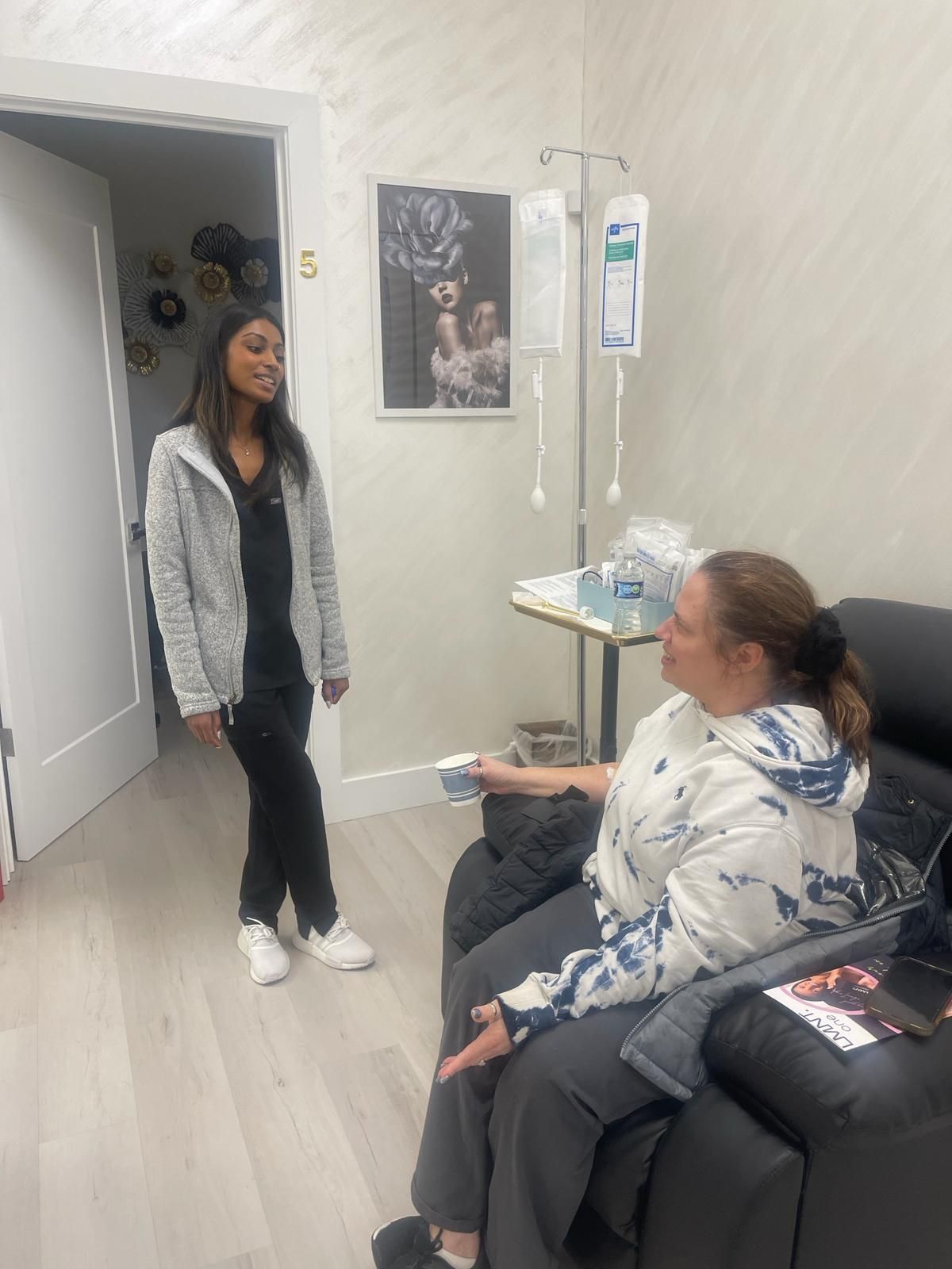 Two people talking in a clinic waiting room, one standing by a doorway and one seated on a couch.
