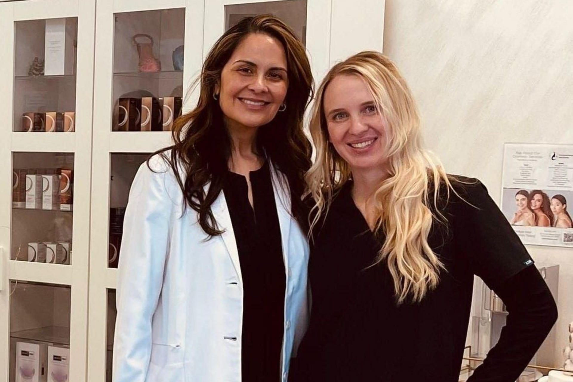 Two women standing in a bright salon, smiling, one in a white coat and one in black clothes